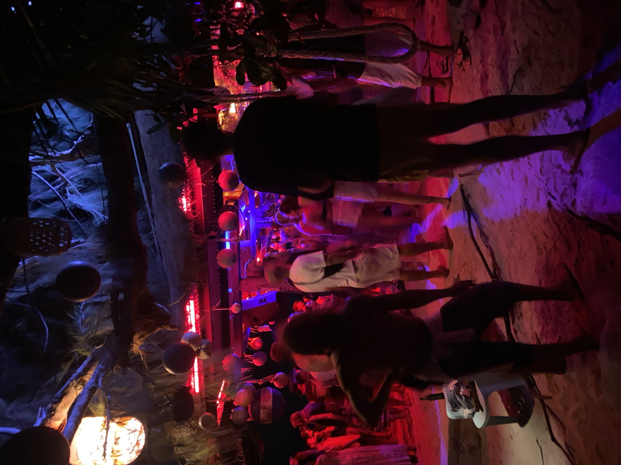 The image depicts a nightlife scene in Baan Sri Thanu, Thailand, featuring a group of people gathered in a space with distinctive decor. The scene is illuminated with red and blue lighting, creating a vibrant atmosphere. People are standing and interacting; their clothing includes shorts and t-shirts. The environment has a beach-like surface, possibly sand, and is decorated with hanging spherical objects. There is a Chang beer logo visible, suggesting the presence of a bar or establishment. The decorations create a lively and possibly tropical or nautical-themed setting.