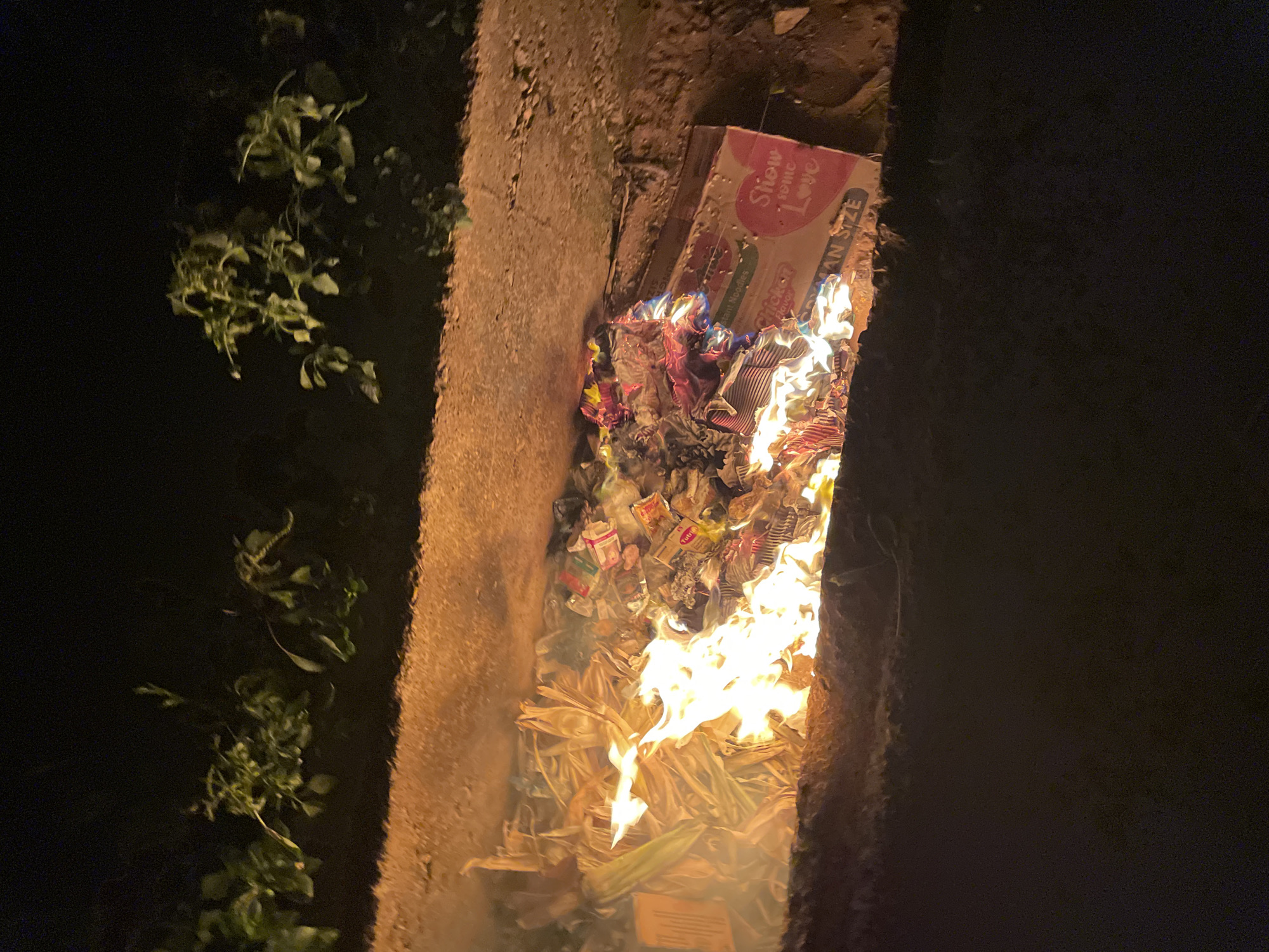 The image shows a fire burning within a narrow, concrete-lined ditch. The flames are consuming what appears to be a mix of paper, cardboard, and other combustible materials. A cardboard box with the words "Show Some Love" is partially visible at the top of the burning pile. Vegetation is visible on the left side of the ditch. The environment is dark, suggesting it is nighttime, and the image was taken in Akwanga, Nigeria.