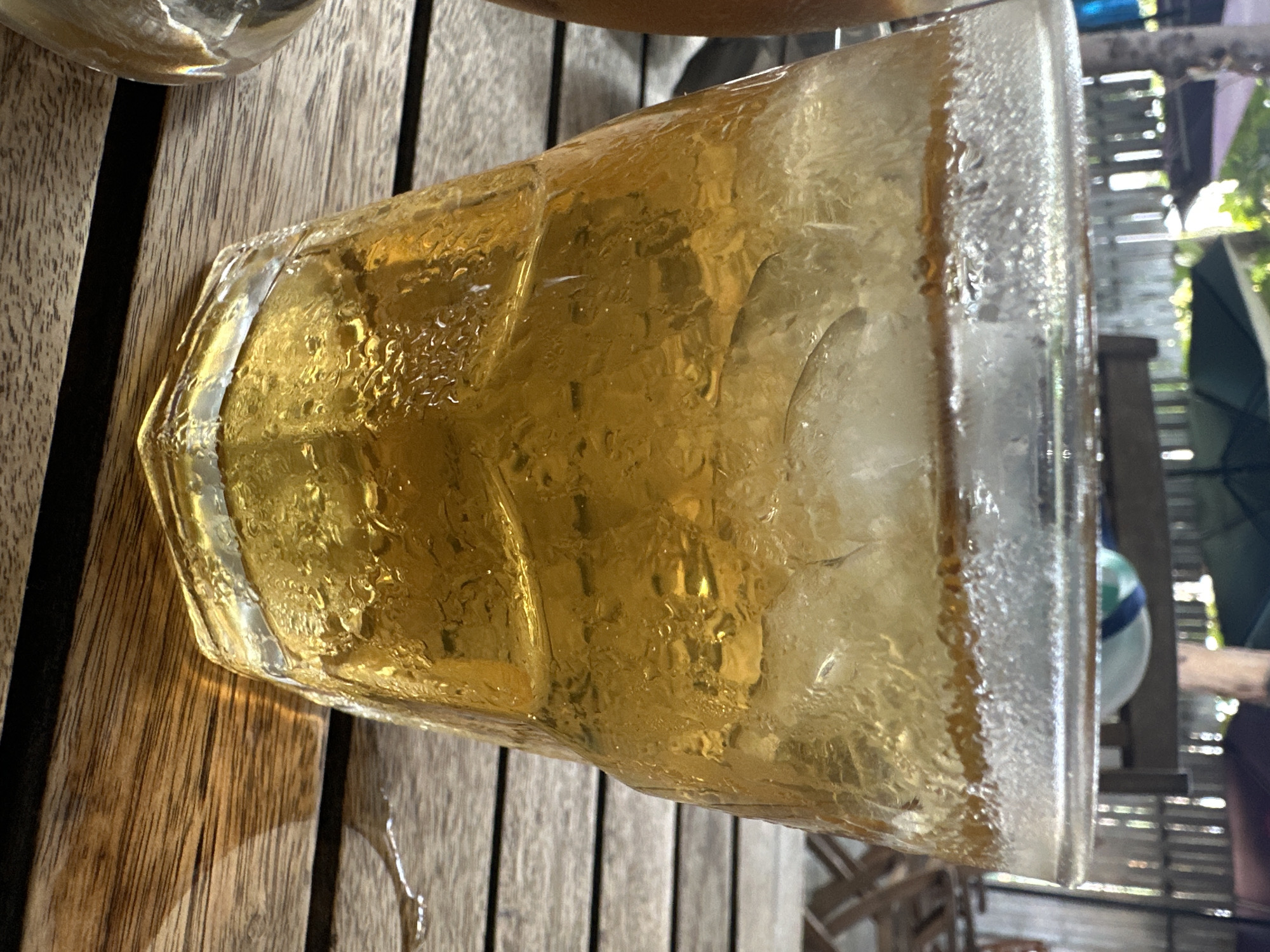 A glass of iced tea sits on a wooden table in what appears to be an outdoor setting. Condensation covers the glass, suggesting the drink is cold. Outside the immediate area, a patio scene is visible with chairs and glimpses of foliage. The location is identified as Nha Trang Ward, Vietnam.