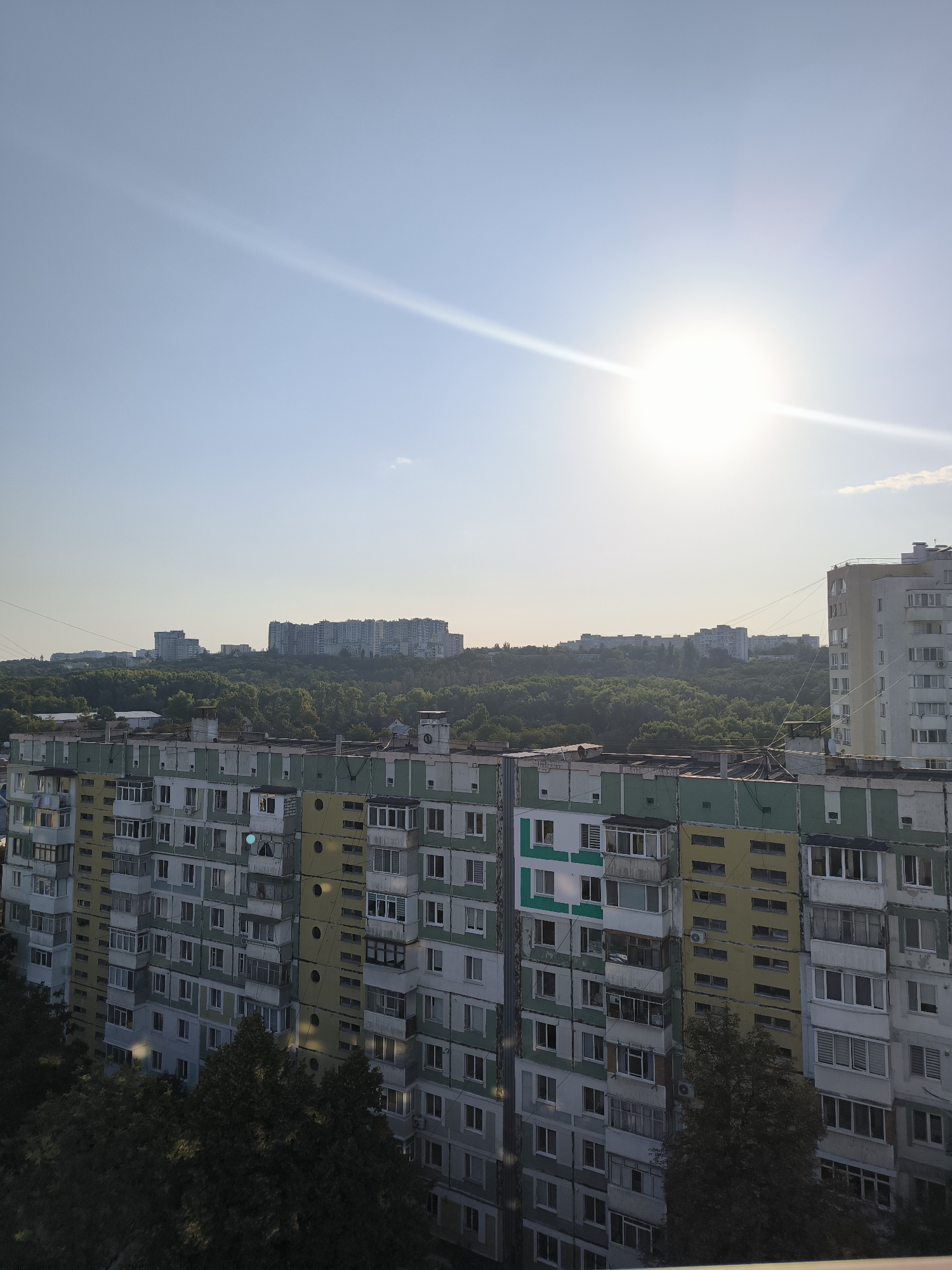The image depicts a wide shot of a residential area in Chișinău, Moldova, likely taken from a high vantage point. The foreground is dominated by the facade of a multi-story apartment building, characterized by its colorful exterior with sections painted in green, yellow, and gray. The building features numerous windows, balconies, and what appear to be air conditioning units. In front of this building, dark green trees are visible, partially obscuring the lower floors.
In the middle ground, another, taller apartment building is visible on the right side of the frame, with a lighter, predominantly white facade. Beyond this, a dense forest covers a hilly landscape, dotted with more apartment buildings of varying sizes and architectural styles peeking through the trees. The sky is clear and bright blue, with the sun prominently featured in the upper right quadrant, casting a strong glare and lens flare across the image. The overall impression is a view of urban sprawl interspersed with natural greenery, under a bright sunny sky.