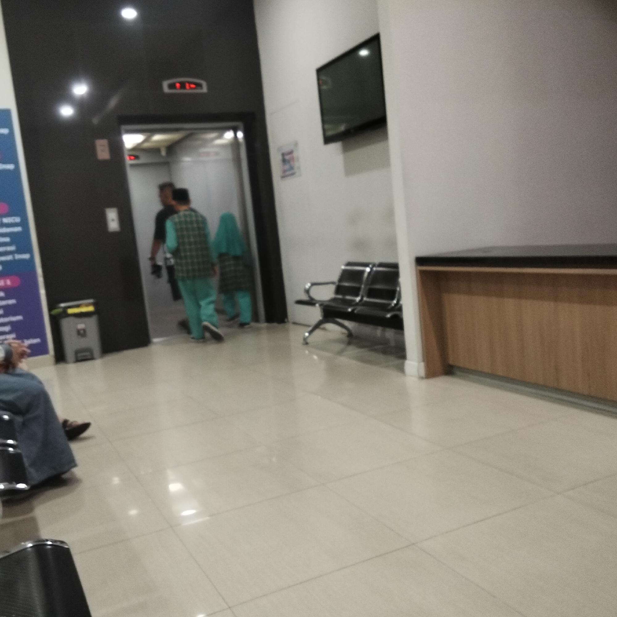 The image depicts an interior scene, possibly a waiting area or hallway within a building, with a focus on a hallway leading to an elevator. On the left, a dark-colored wall features a blue and purple informational sign with white text, partially obscured. Adjacent to the sign is a grey trash receptacle. Further to the right, an elevator entrance is visible, with two individuals near the entrance. One person is seen from the back, wearing a green and black checkered vest over a light green outfit, and appears to be walking away from the elevator. Another person, wearing a teal head covering and a teal garment, is seen from the back, standing near the elevator. Red indicator lights are visible above the elevator doors. In the background, to the right of the elevator, a dark flat-screen television is mounted on a white wall. Below the television, a long, light-brown reception desk with a dark top extends across the wall. In the mid-ground, a black metal bench with two seats is positioned on the tiled floor. In the foreground, on the left, a person is seated, wearing a dark blue garment. The floor throughout the scene is covered in large, light-colored tiles. The lighting appears to be artificial, with several overhead lights visible. There are no clear indicators of the specific city of Sukabumi, Indonesia, within the image itself, though the context provided by the prompt implies this location.