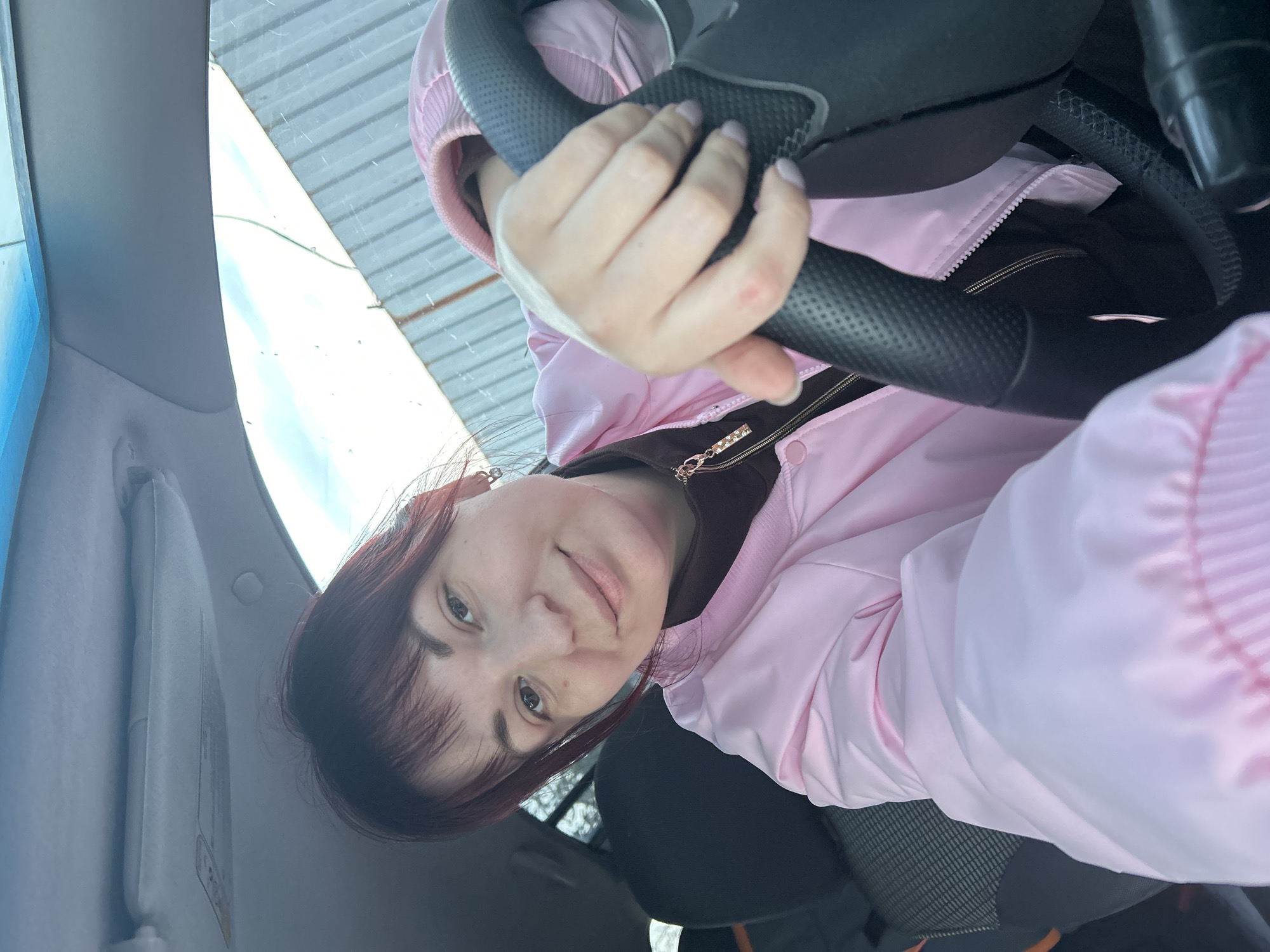 A woman is in the driver's seat of a car. She is holding the steering wheel with both hands. She is wearing a pink jacket over a brown top with a gold zipper. The car's visor is visible. Through the windshield, there is a blurry view of what appears to be a corrugated metal roof or fence. The location is presumed to be in the city of Фоки, Russia, but this cannot be confirmed visually from the image.