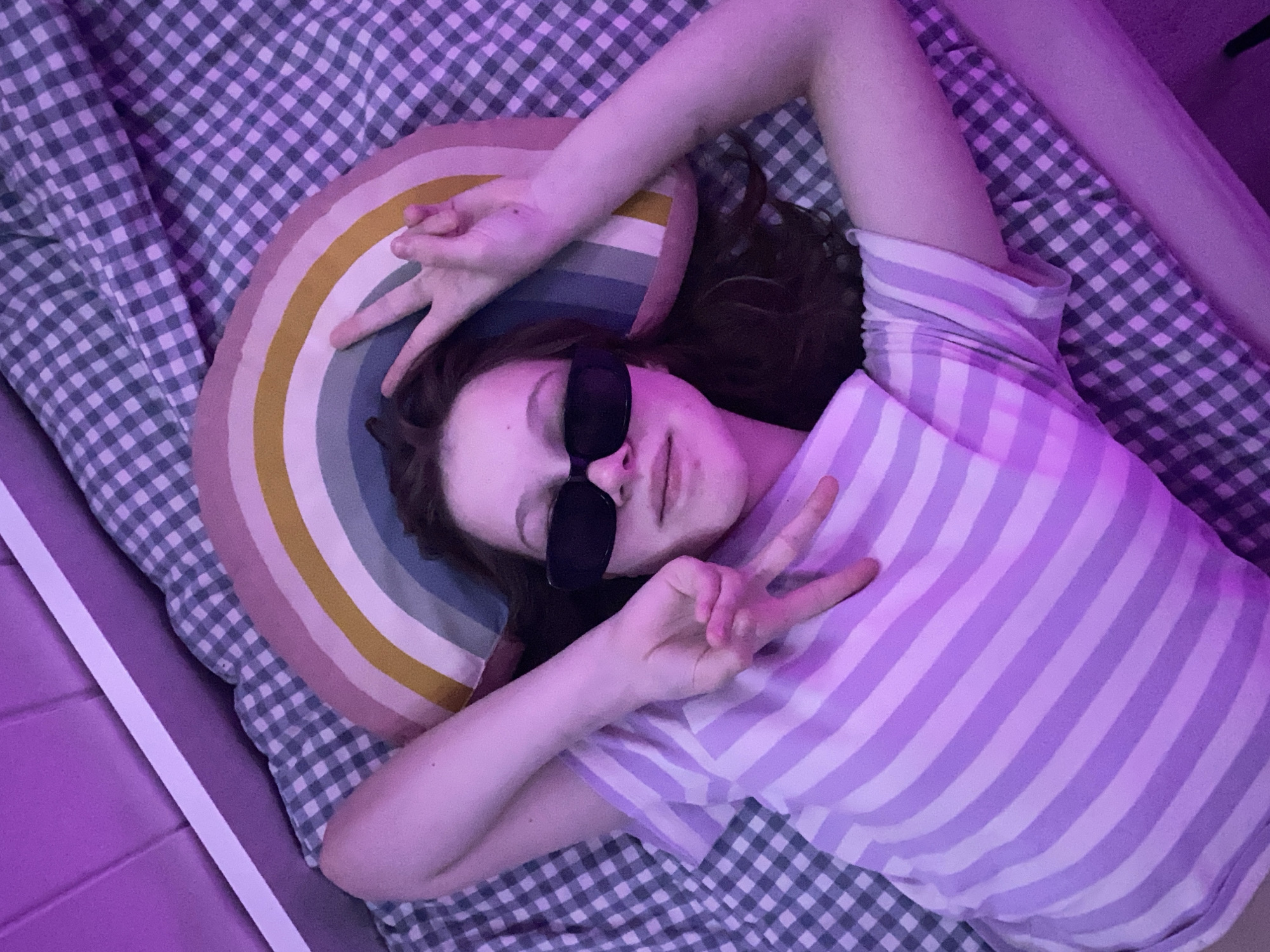 A person wearing sunglasses and a striped shirt is lying on a checkered bedspread. They are positioned on top of a rainbow-shaped pillow and making peace signs with both hands. The scene is illuminated by purple lighting, and there is a white border of a bed frame visible in the lower-left and upper-right corners. The location is likely Chaikovsky, Russia.