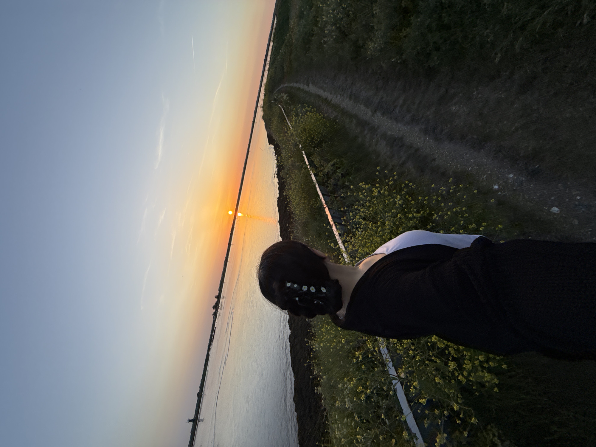 The image shows a person from the back, wearing a dark top over a white shirt, with a dark hairstyle decorated with small white flower accessories, standing by a waterway at sunset. To the left is the waterway reflecting the orange and yellow hues of the setting sun, with a clear sky transitioning from blue to orange. On the right is a grassy bank covered in yellow wildflowers with a visible dirt path alongside. The environment suggests a rural setting near water. The location could possibly be in the city of Great Yarmouth, United Kingdom, but this is speculation.