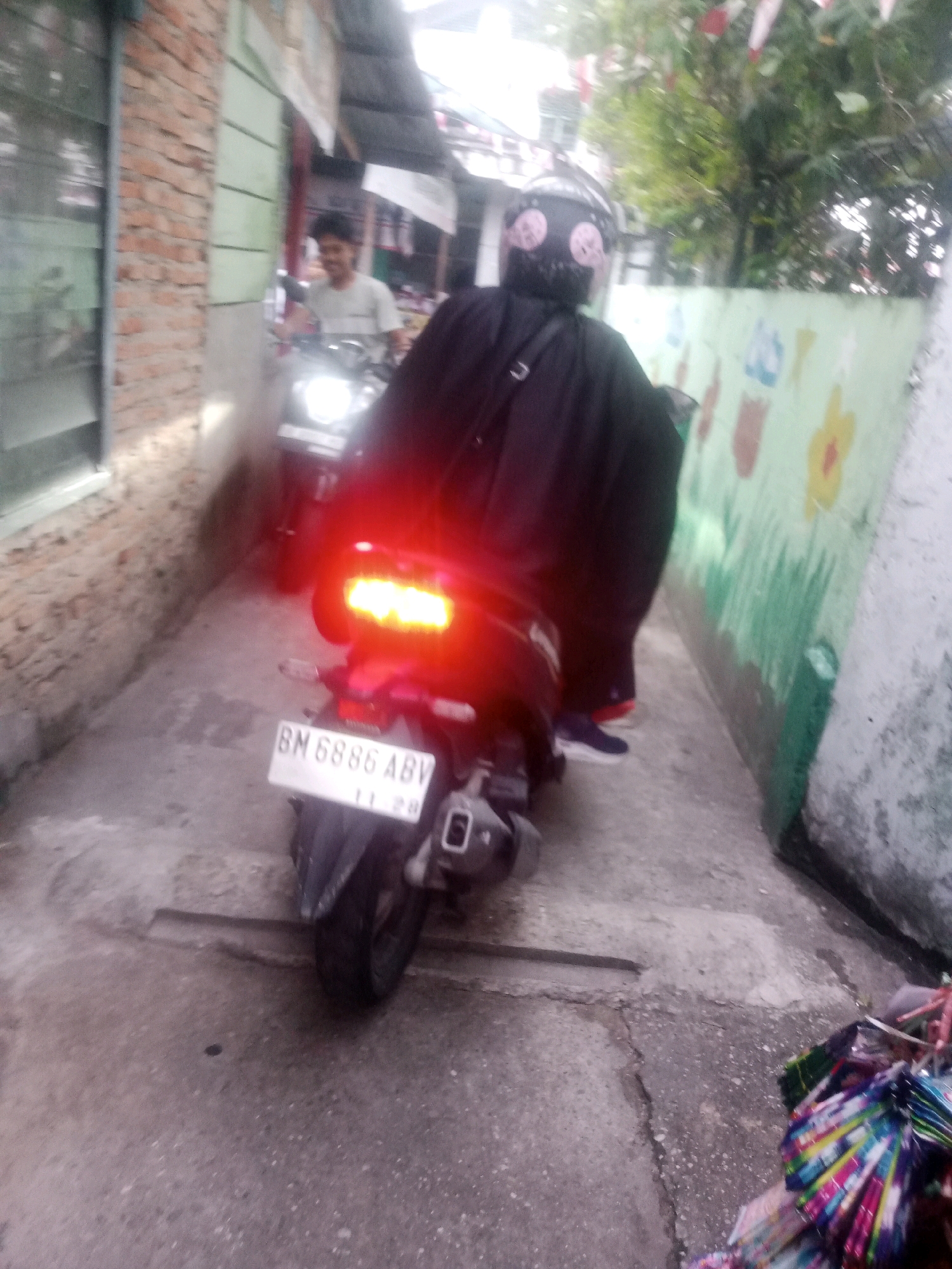 The image depicts a narrow street in Pekanbaru, Indonesia, with a person riding a motorcycle. The rider is wearing a black cloak and a helmet with a skull design. The motorcycle has a license plate that reads "BM 6886 ABV". The street is paved with concrete and lined with buildings on both sides. There is a wall on the right side of the street with colorful drawings. In the background, there are trees and other buildings. The lighting suggests it is daytime.