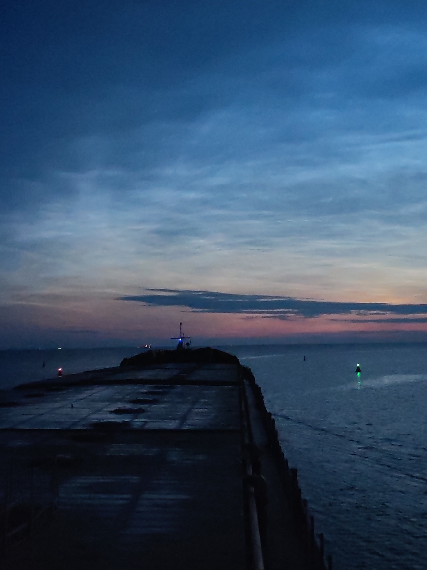 At dusk, a view of a harbor in Ozernoe Ust'e, Russia, shows a concrete pier jutting into calm, dark water. The sky above is a gradient of deep blue, transitioning to lighter shades of blue and streaks of pink and orange near the horizon. A few thin clouds are visible in the upper sky. On the pier, a structure topped with a blue light is illuminated. Further out in the water, a green navigation buoy glows, marking a channel. The pier's surface appears wet and reflective in places, with visible seams and some darker, stained areas. The overall atmosphere is tranquil and indicative of the transition from day to night.