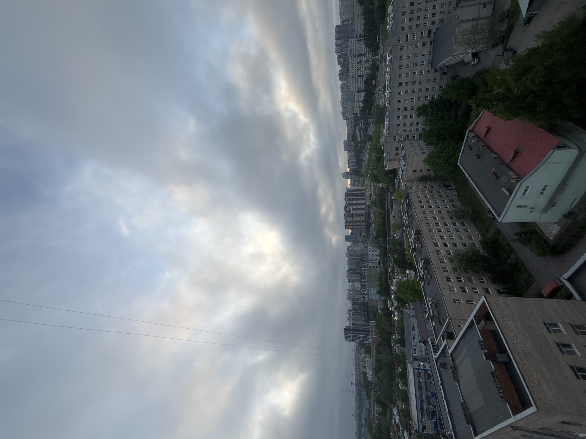 The image depicts an urban landscape viewed from an elevated position, possibly a high-rise building. The city comprises a mix of residential buildings, ranging from older, low-rise structures to modern high-rise apartments. Numerous trees line the streets, suggesting a significant amount of greenery integrated into the city. The sky is overcast with heavy clouds. A pair of electrical wires are seen in the lower left of the frame. The buildings are primarily constructed of concrete, with a variety of architectural styles. Cars are parked along the streets, indicating normal urban activity.
