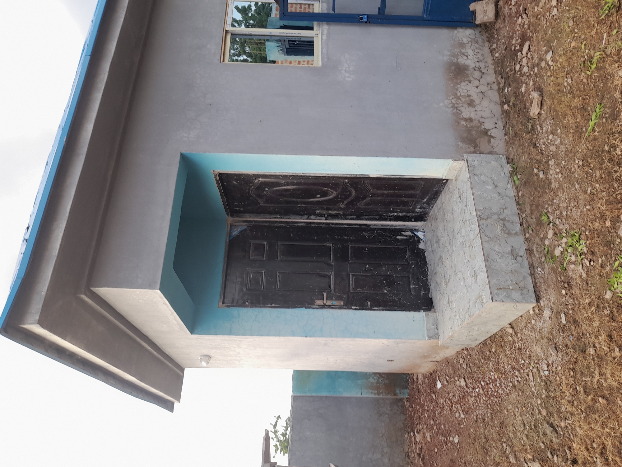 The image shows a small, unfinished concrete building with a blue corrugated metal roof. The building has two black doors side-by-side at the entrance, which is accessed by a small concrete step. There is a window on the right side of the building. The exterior walls are painted gray, with a light blue accent around the doorway. The surrounding area is undeveloped land with dirt, gravel, and some sparse vegetation. There is a concrete wall in the background, painted with blue accents. The scene appears to be in the city of Ibi, Nigeria, based on the context provided. There are no people visible in the image.