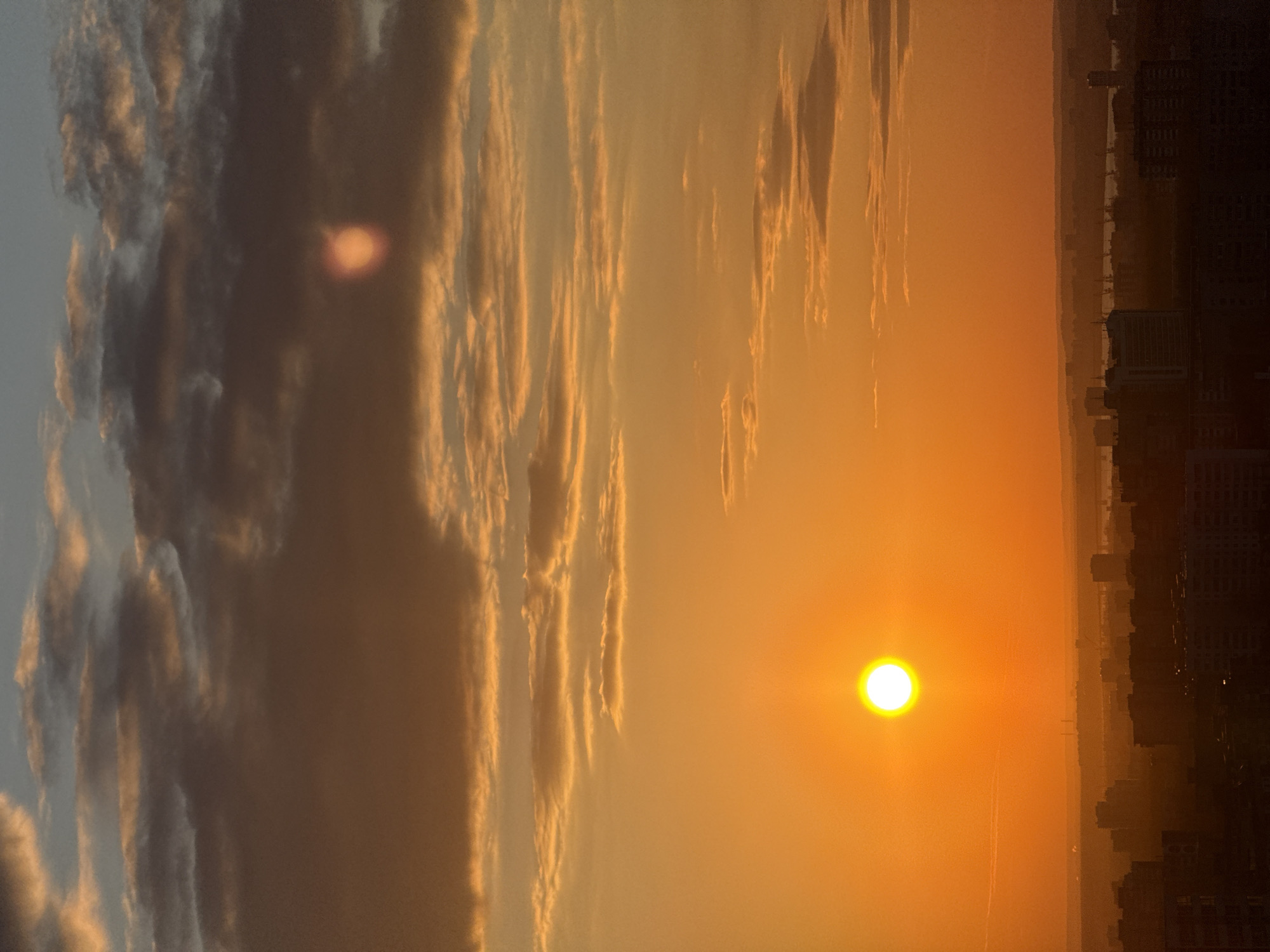 This photograph captures a vibrant sunrise or sunset over an urban landscape in Perm, Russia. The sky dominates the scene, painted in rich hues of orange and yellow, with a bright, glowing sun positioned prominently in the mid-right. Streaks of clouds, some dark and voluminous, others illuminated with warm golden edges, stretch across the sky, creating a dynamic visual texture. The atmosphere appears hazy, contributing to the soft, diffused light and warm color palette. Along the bottom edge of the image, a distant city skyline is silhouetted against the bright horizon, indicating an urban setting. Further beyond the city, faint layers of hills or distant land are visible through the haze. No human activity or visible text is present. The golden hour light creates a serene and picturesque mood, characteristic of either daybreak or dusk over the city.