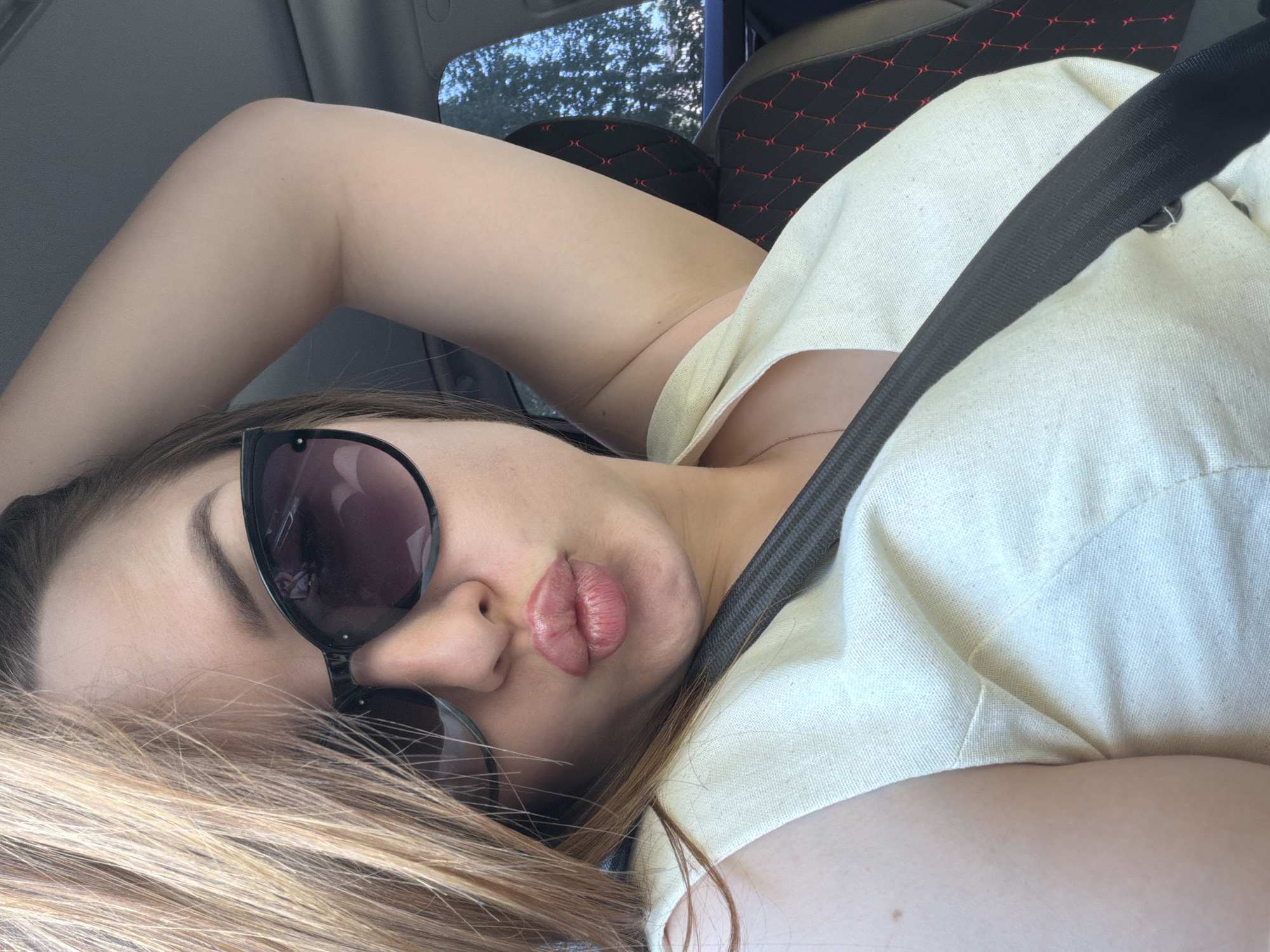 A woman with fair skin and light brown hair is wearing a seatbelt and sunglasses while lying down in a car. She is wearing a light-colored shirt and has her arm raised. The car has black seats with a red diamond pattern. The image may have been taken in daylight. There is no indication of the location being in Chaikovsky, Russia.