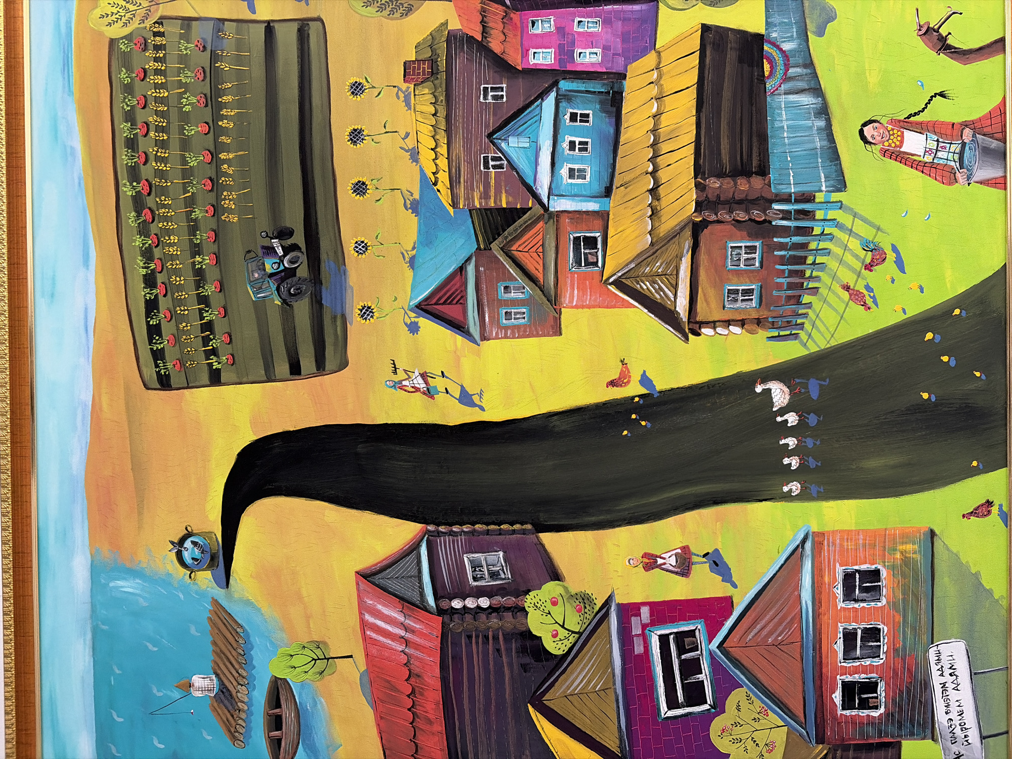 A painted canvas depicts a bird's-eye view of a rural scene, enclosed within a golden frame. The scene consists of structures and agricultural elements that suggest a small village or rural community. The painting style is naive and employs vibrant colors.
At the top left, a rectangular plot of land features rows of crops and a tractor, suggesting agricultural activity. Beside it, there is a body of water with a person fishing from a wooden raft. A boat is visible nearby.
A black winding road or path leads to a village with houses. The structures display different colors. Figures of people are interspersed between the houses. A woman is depicted near the upper right corner. Birds and other small animals are scattered across the canvas.
The lower right of the painting contains text or inscriptions written on what seems to be a wall. The absence of recognizable landmarks precludes confirmation of location.