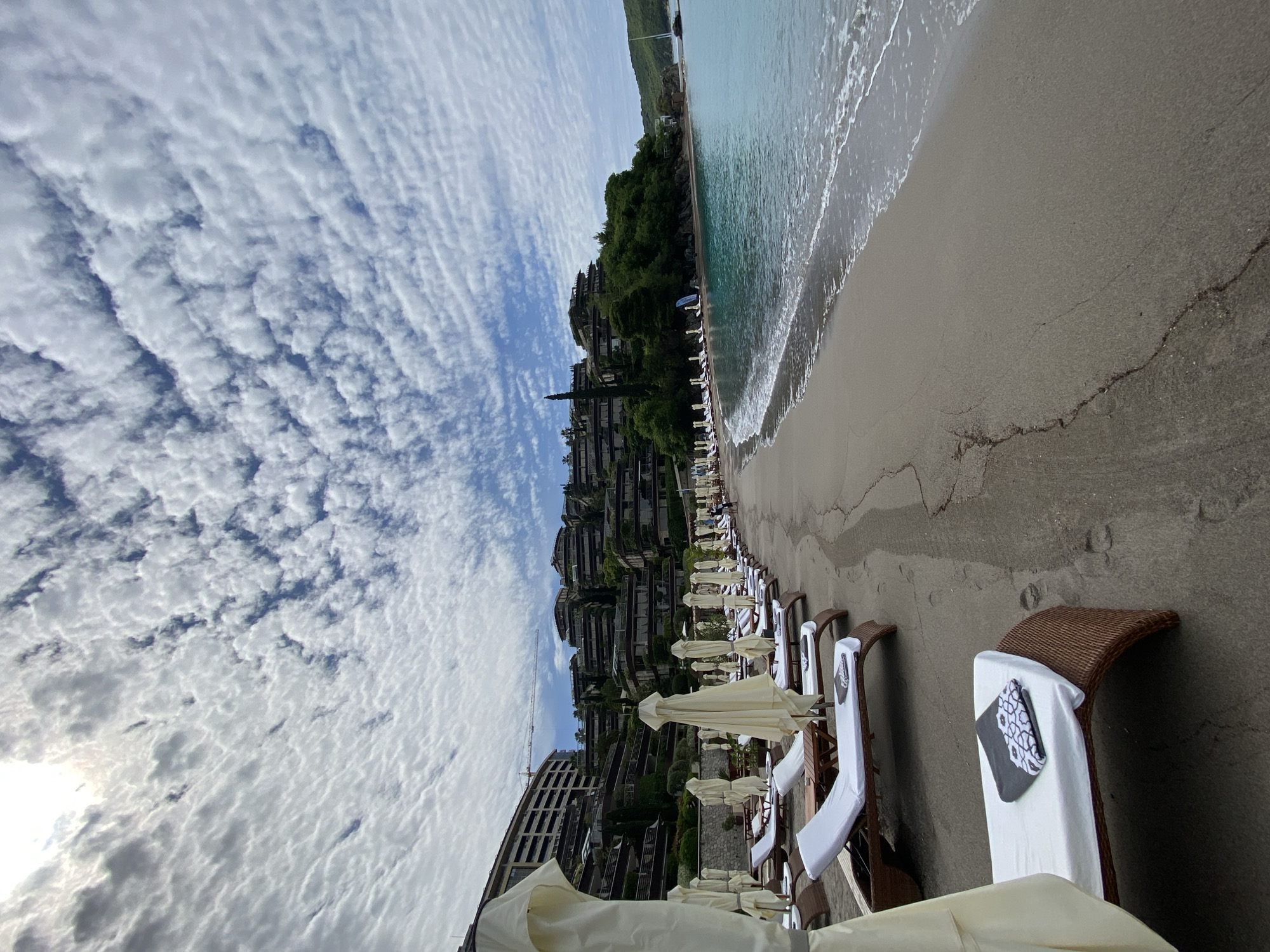 The image depicts a beach scene in Budva, Montenegro, with a focus on the beachfront amenities and the surrounding environment.
In the foreground, numerous lounge chairs with white towels are arranged in rows, most with closed cream-colored umbrellas. The chairs are of a dark brown wicker design. A black and white patterned object, possibly a book or magazine, is visible on one of the chairs.
The beach itself is sandy with a dark grey hue and visible cracks. The water's edge is marked by small waves. Behind the beach, a hill is covered with dense greenery and interspersed with multi-story buildings that cascade down the slope. These buildings feature staggered balconies and are likely part of a resort or residential complex.
The sky is filled with a layer of scattered cumulus clouds against a blue sky. Overall, the scene suggests a calm day at a well-maintained beach resort.
