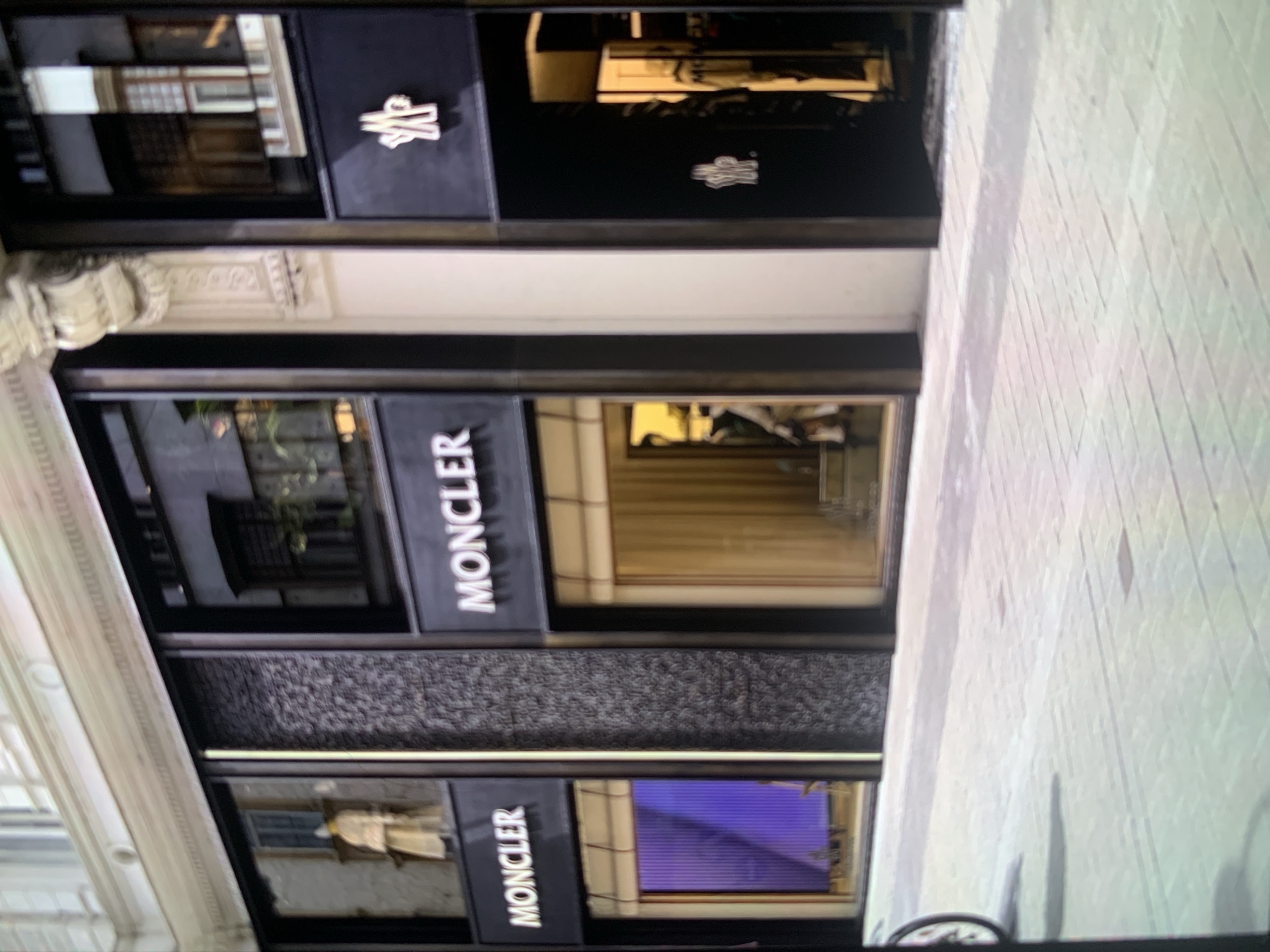 The image shows the exterior of a Moncler retail store located on an urban street. The storefront features a modern, sleek design with prominent black panels and large glass display windows. The brand name "MONCLER" is clearly visible in white capital letters on multiple black signs integrated into the facade. Above one of the display windows, the distinctive white Moncler logo, a stylized monogram, is also visible on a black background.
The store is housed within a building that exhibits classical architectural details, including ornate light-colored stone moldings and cornices along its upper sections and sides, creating a contrast with the contemporary store design. Reflections in the store windows show glimpses of other buildings and the street scene opposite. In one of the lower windows, a vibrant purple light or display is visible within the store interior.
The ground area in front of the store appears to be a light-colored paved sidewalk or street with a grid pattern. No people or specific activities are depicted in the scene, which appears to be a static shot of the storefront during daytime.