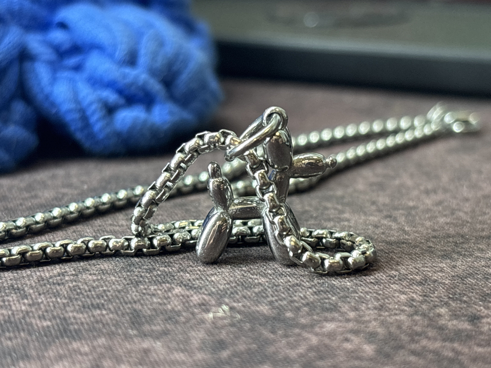 The image depicts a silver necklace with a balloon animal pendant on a textured brown surface. A crumpled blue fabric is visible in the background to the left, and a dark, possibly electronic, object is in the upper right. The scene lacks contextual information to place it specifically in Perm, Russia.