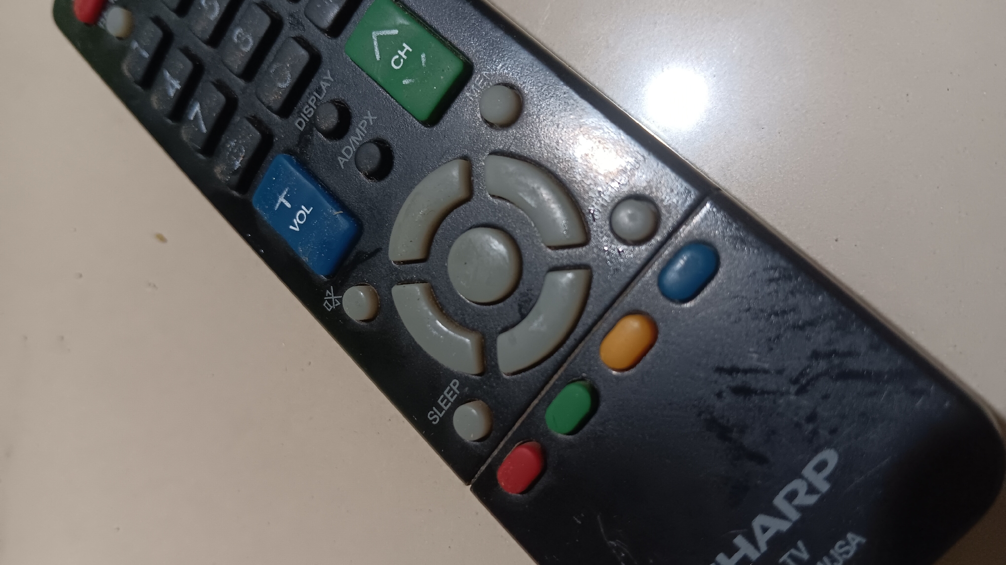 The image shows a black Sharp TV remote control lying on a light-colored, textured surface, possibly a table or countertop. The remote is angled, with the top right corner closer to the viewer. Several buttons are visible, including a blue button with "VOL" printed on it, a green "CH" button, and a set of directional arrows surrounding a central button. There are also red, green, orange, and blue oval buttons at the bottom left of the remote. Some of the buttons and the remote's surface appear dusty or smudged. The Sharp logo is visible on the lower left portion of the remote. There are no people or any indication of a specific location within the city of Sonobekel, Indonesia. The environment appears to be indoors, with artificial lighting that causes a bright reflection on the surface to the right of the remote.