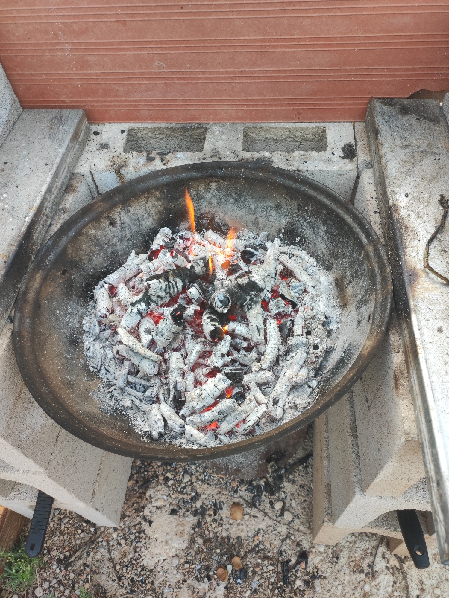 The image shows a close-up, overhead view of a metal fire pit filled with glowing embers and ash. The fire pit is supported by concrete blocks, with a red, ribbed surface above it, possibly a wall or overhang. The ground beneath is covered in gravel, small stones, and some scattered ash. There are no people or animals visible in the image. The scene appears to be outdoors, possibly in a backyard or patio area. The location is identified as Alcàsser, Spain.
