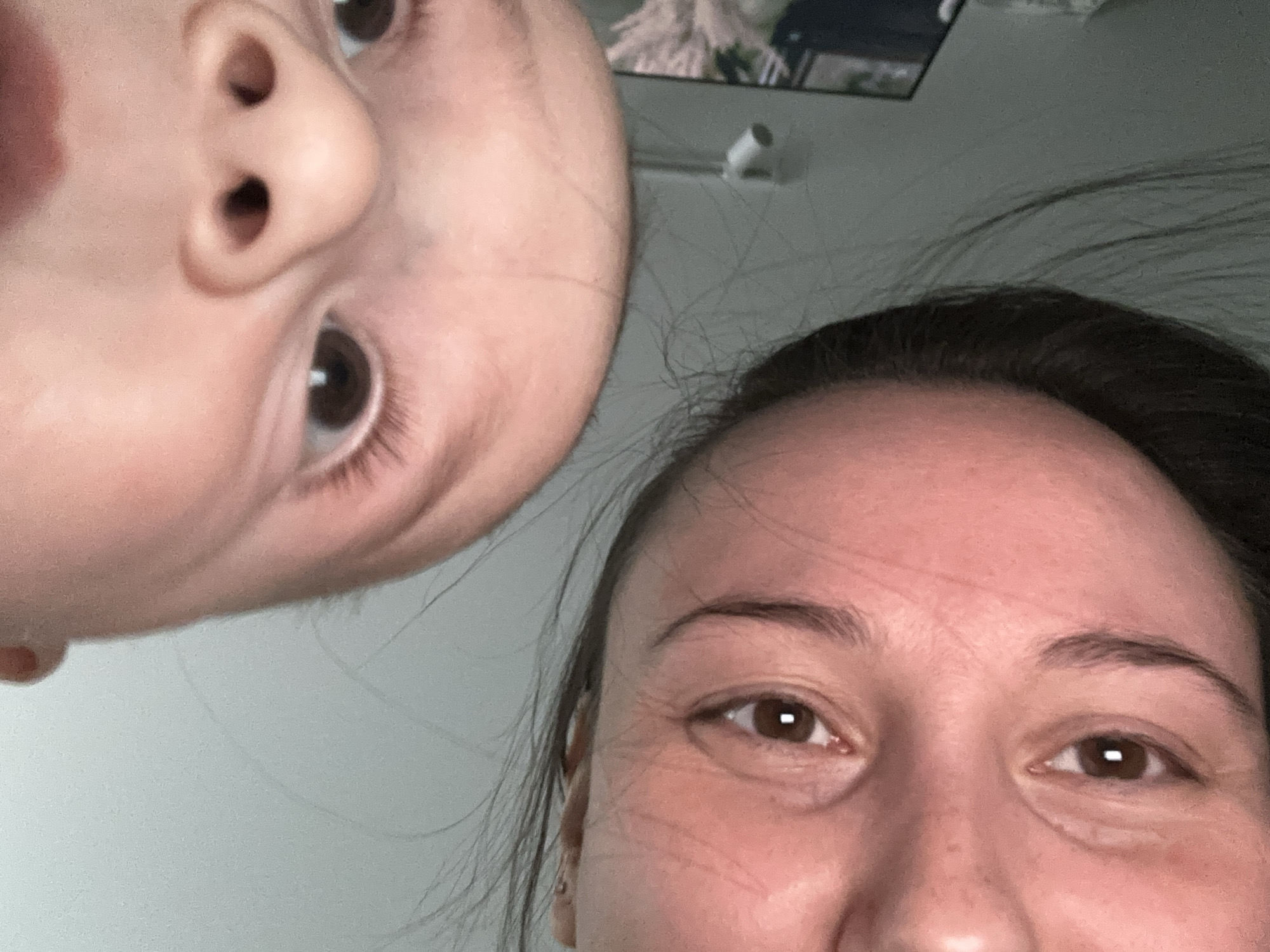 The image presents a close-up, inverted view of two individuals in an indoor setting within Chaikovsky, Russia. The lower left portion of the frame features the upside-down face of an adult female with dark hair, visible brown eyes, and dark eyebrows, looking towards the camera. The upper right portion of the frame contains the upside-down face of a younger child, likely an infant or toddler, also with brown eyes and a prominent, small nose, similarly looking towards the camera. The background consists of a plain, light-colored wall or ceiling. A blurry reflection of a decorative object and a small white wall fixture are faintly discernible in the background towards the lower right. The specific location within Chaikovsky is not visually identifiable from the image content.