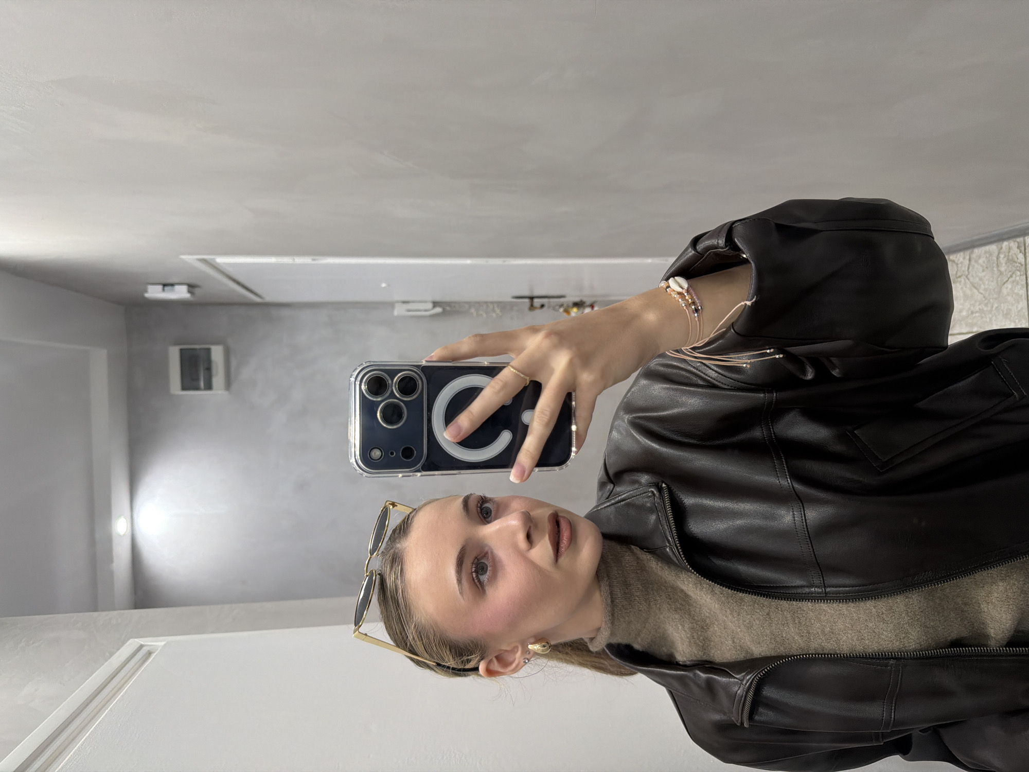 A young adult female is depicted in a mirror selfie, located in an indoor setting in Plovdiv, Bulgaria. She is facing slightly to her right, holding a dark-colored smartphone with a clear MagSafe case in her right hand, capturing her reflection. Her light brown hair is pulled back, with gold-rimmed oval sunglasses resting on top of her head. She wears a gold hoop earring on her visible left ear, and a gold ring on her left ring finger. Her makeup includes noticeable pink blush on her cheeks and light brown lipstick. She is dressed in a brown turtleneck sweater under a dark brown leather jacket, partially unzipped. Her right wrist displays multiple bracelets, including one with small beads and a cowrie shell, and another with thin pink strings and silver beads. A dark-colored leather purse is carried on her right arm. The background, visible in the reflection, features light grey walls with a textured finish. A white door frame is visible on the left, and a white door is further right. A recessed ceiling light fixture is reflected, emitting bright illumination. An electrical panel or fuse box is mounted on the wall to the left. The floor appears to be light-colored tile.