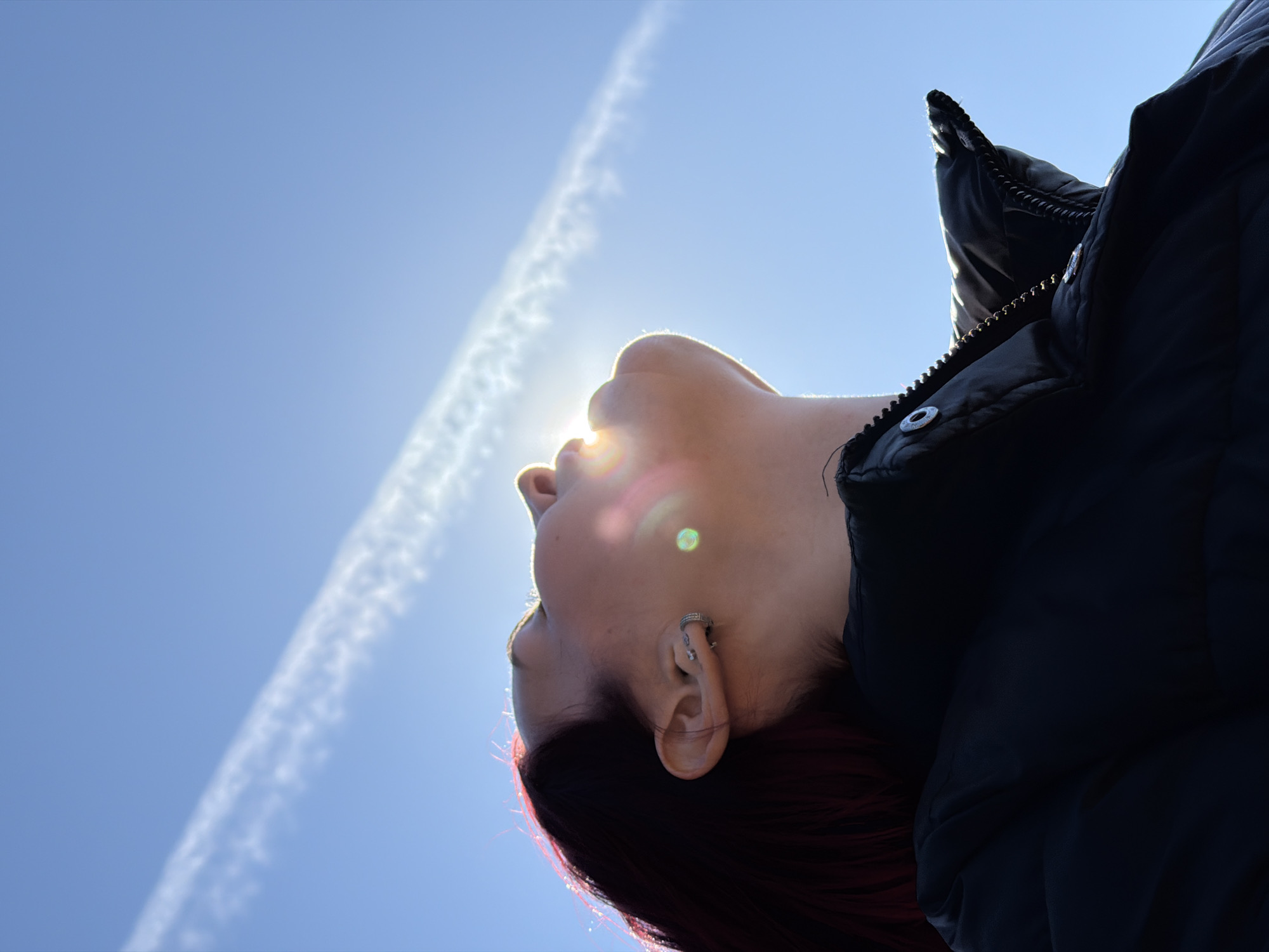A low-angle shot depicts a person with red-tinted hair, wearing a dark, possibly black, jacket, tilting their head upwards towards the sun. The sun is positioned directly in front of their face, causing a bright flare. A contrail cuts across the blue sky in the background. The person is wearing a small earring in their left ear. The location is not evident from this image. The city of Chaikovsky, Russia, cannot be determined from the image.