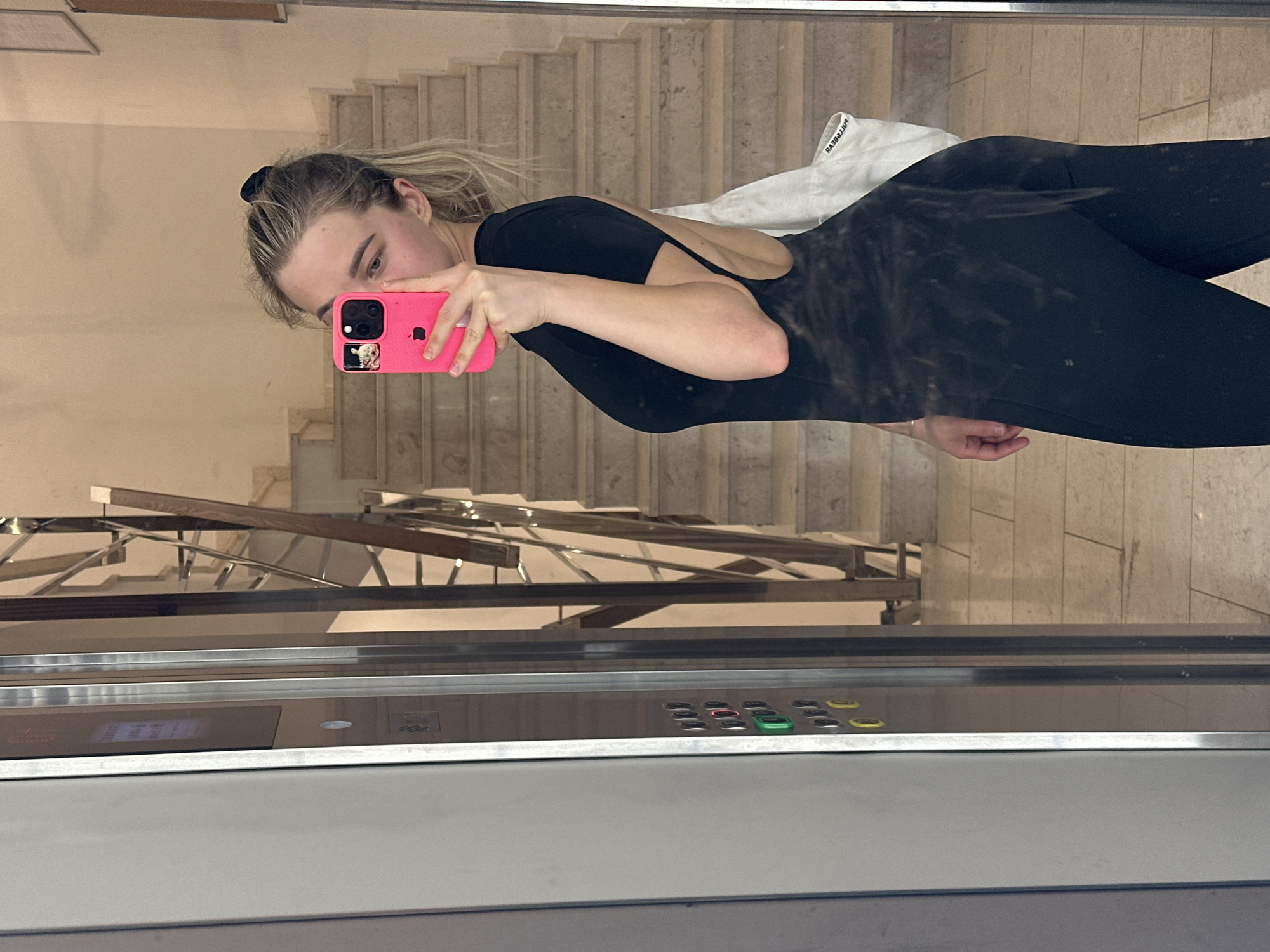 A young woman with blonde hair, partially tied back with a black scrunchie, is taking a mirror selfie in what appears to be an elevator in Budva, Montenegro. She is holding a bright pink iPhone, which covers most of her face except for her right eye and eyebrow. On the phone's screen, a small image, possibly of a cat, is visible. The woman is wearing a fitted black jumpsuit with an open back. A white piece of fabric, possibly a shirt, with reversed text "BIRREBE..." is seen over her left shoulder.
The background, visible through the reflective surface, features a light-colored staircase with metal railings, suggesting an indoor setting like a hotel or residential building. The lower part of the image shows an elevator control panel with various buttons, including prominent green, red, and yellow ones, and some faint, illuminated text or symbols. The overall scene is well-lit, indicating an indoor environment.