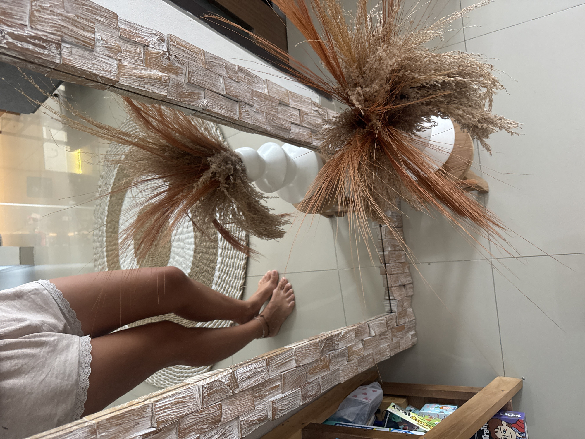 The image shows a reflection in a mirror with a textured, wooden frame. The reflection captures a person's legs and feet, clad in light-colored shorts with a lace trim. The legs are positioned against a background of a woven mat or rug. In the foreground, positioned partially in front of and beside the mirror, is a floral arrangement with dried grasses and reeds in a white, bulbous vase. The surrounding environment consists of tiled walls. Below the mirror, there is a wooden structure filled with various objects, including boxes and books. The location of the scene is an interior space. The city and location are unspecified.
