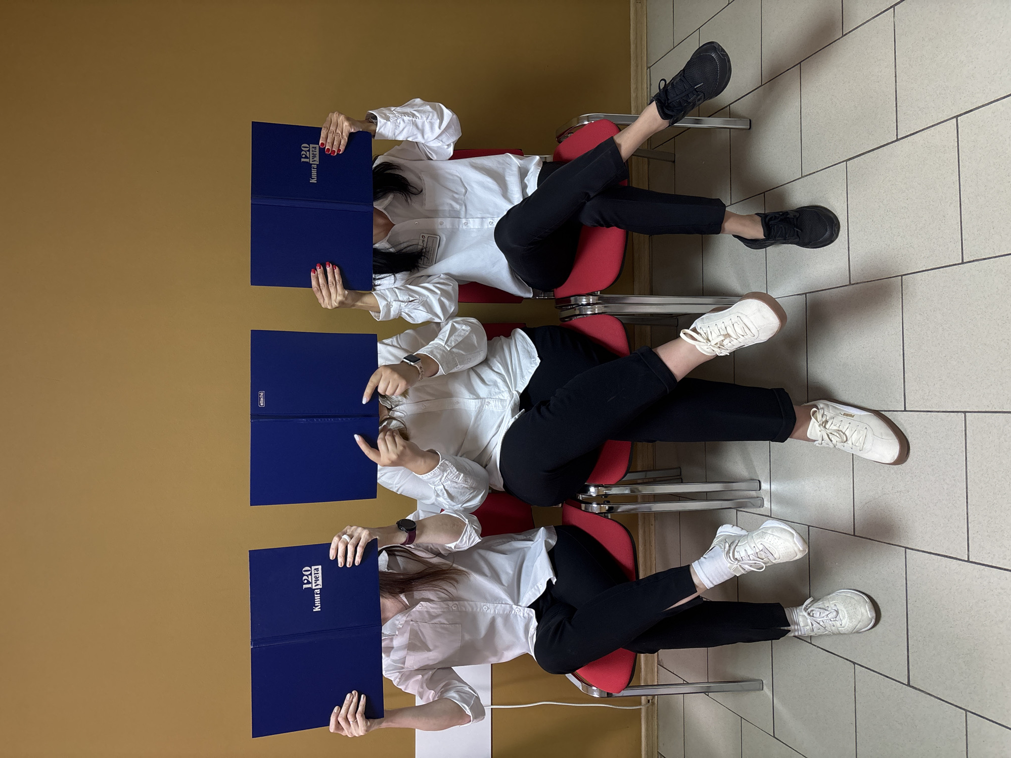 Three women are seated in red chairs in a tiled hallway against a mustard-colored wall. Each woman wears a white, button-down shirt and black pants. They are holding open blue books in front of their faces. The book covers display text in Russian. The women wear different styles of white and black sneakers. The chairs are stacked slightly behind each other, in a row. This photograph was taken in the city of Chaikovsky, Russia.