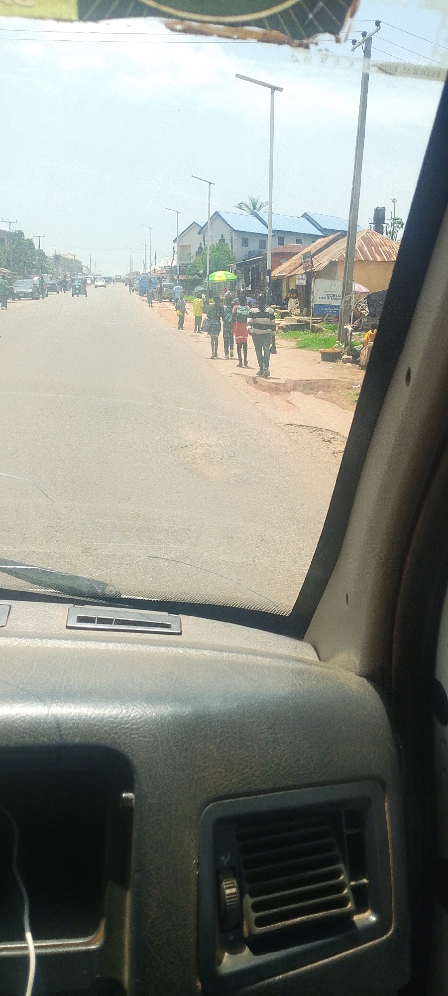The image captures a street scene in Jos, Nigeria, viewed from inside a vehicle. The sun is high in the sky, indicating it is likely daytime, and the weather appears clear and bright, casting distinct shadows.
The main subjects are several people walking along the side of a paved road. Some individuals are carrying items, suggesting they might be pedestrians on their way to or from a market or their homes. There are also vehicles on the road, including cars and what appears to be a three-wheeled auto-rickshaw, typical of transportation in the region. Buildings line the street, some with corrugated metal roofs, and others appear to be more modern with blue roofs. Streetlights are visible along the road.
The scene is a bustling street, likely a commercial or residential area, with people engaged in their daily activities. The overall mood seems ordinary and active, reflecting the typical movement of a city. There are no strong emotional cues to discern from the people depicted.
Notable details include a cracked windshield visible in the foreground, suggesting the vantage point is from within a car with some wear. The environment appears somewhat dusty, a common characteristic of urban areas in certain climates.
While most text is illegible due to distance and image quality, a sign on one of the buildings appears to have some writing, possibly business names or information, but it is too blurry to read clearly. Another signpost has text that reads "COCIIN LC..TUODU WADA".