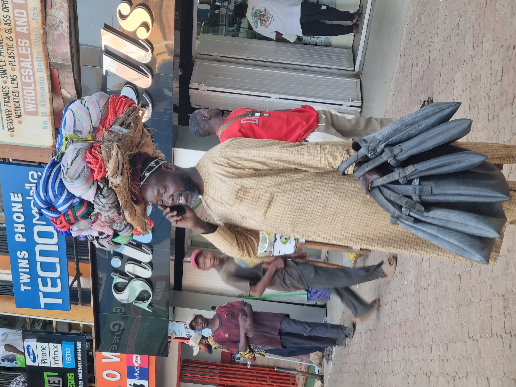 The image captures a bustling street scene in Jos, Nigeria, specifically on Ahmadu Bello Way. A man with a gray beard and wearing a white-patterned cap and tan robe is walking, carrying a large bundle of colorful striped textiles on his head and a group of dark grey bags in his left hand. He is also holding a smartphone to his ear. He is walking past storefronts. The storefronts contain signage for "Twins Phone, TECNO" and "Silks". Further signage shows an ad for different phone brands. Other people are present in the background, walking in the same direction. The ground is paved with interlocking paving stones.