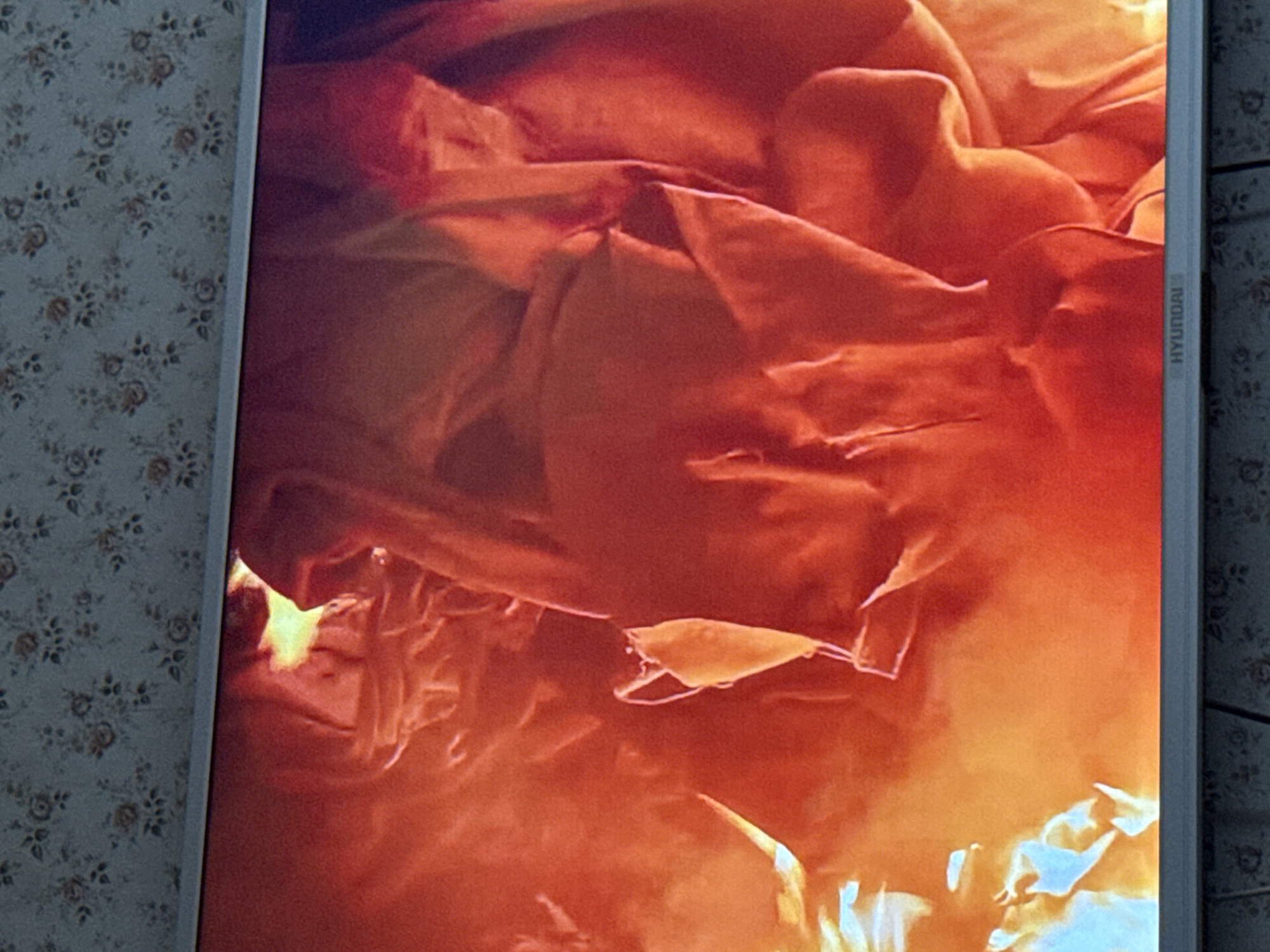 The image appears to be a photograph of a screen displaying an image dominated by orange and red tones, suggesting fire or a similar intense light source. There are shapes that could be interpreted as fabric or other malleable materials caught in the light. The screen's bezel is visible, bearing the brand name "Hyundai". The screen is also positioned against a backdrop of floral wallpaper. There is no indication of a location in the city of Mytishchi, Russia, or of people, subjects, objects, actions, interactions, or the environment beyond what's visible on the screen and its immediate surroundings.