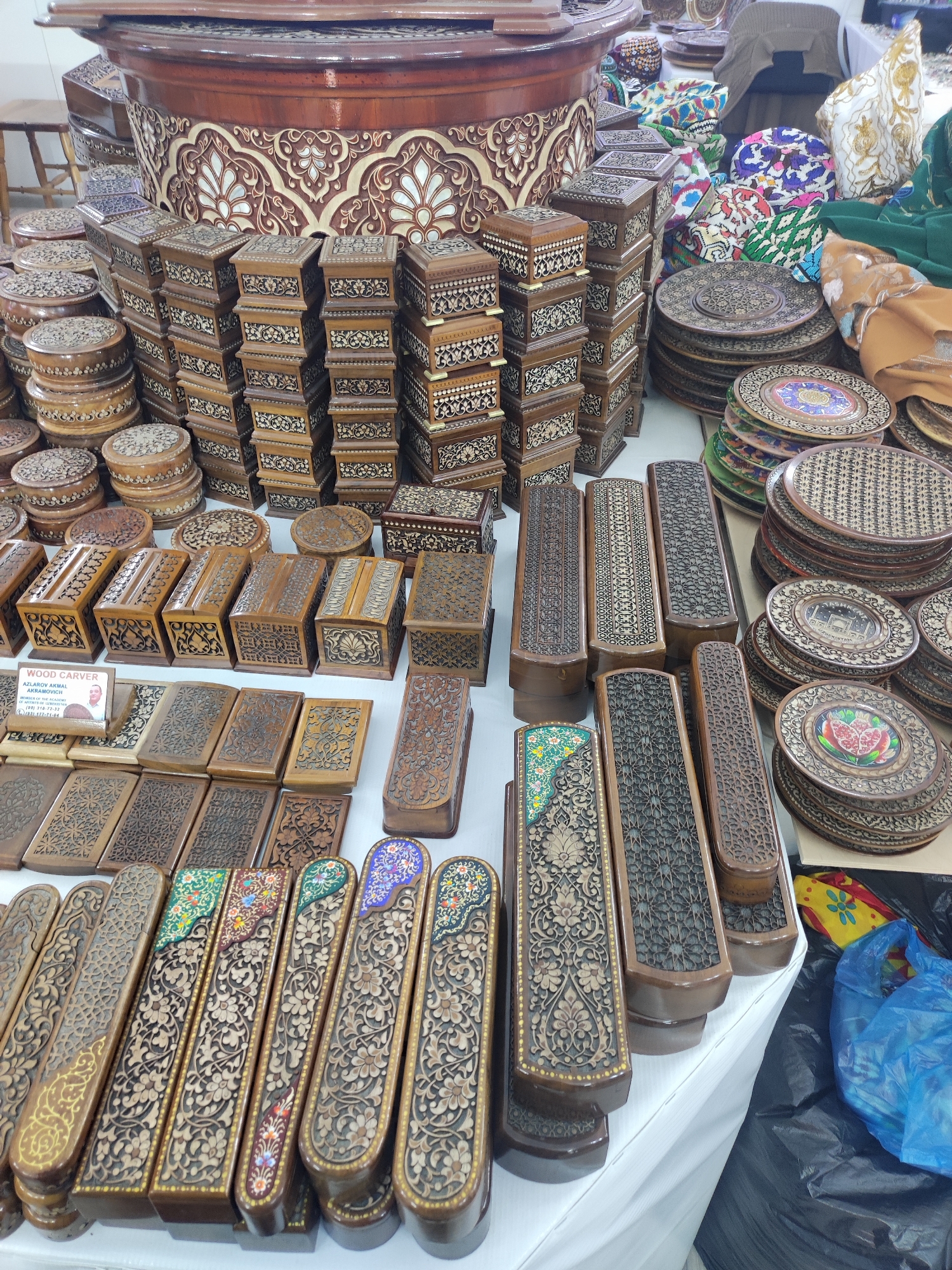 The image shows a market stall with an assortment of wooden items, primarily decorative boxes and plates. The boxes vary in size and design, with intricate carvings and inlaid patterns. Some boxes are stacked in tall columns, while others are displayed individually. There are also round, flat wooden plates with carved designs, some of which feature colorful embellishments. In the background, a large, ornately carved wooden barrel-like structure is visible, along with stacks of patterned fabric items and what appear to be traditional hats. A blue plastic bag and some colorful fabric are on the ground to the right. The lighting suggests an indoor setting, possibly a covered market. No people are clearly visible in the image, though the presence of a stall implies a vendor and potential customers. The overall impression is one of a craft or artisan market selling handcrafted wooden goods.