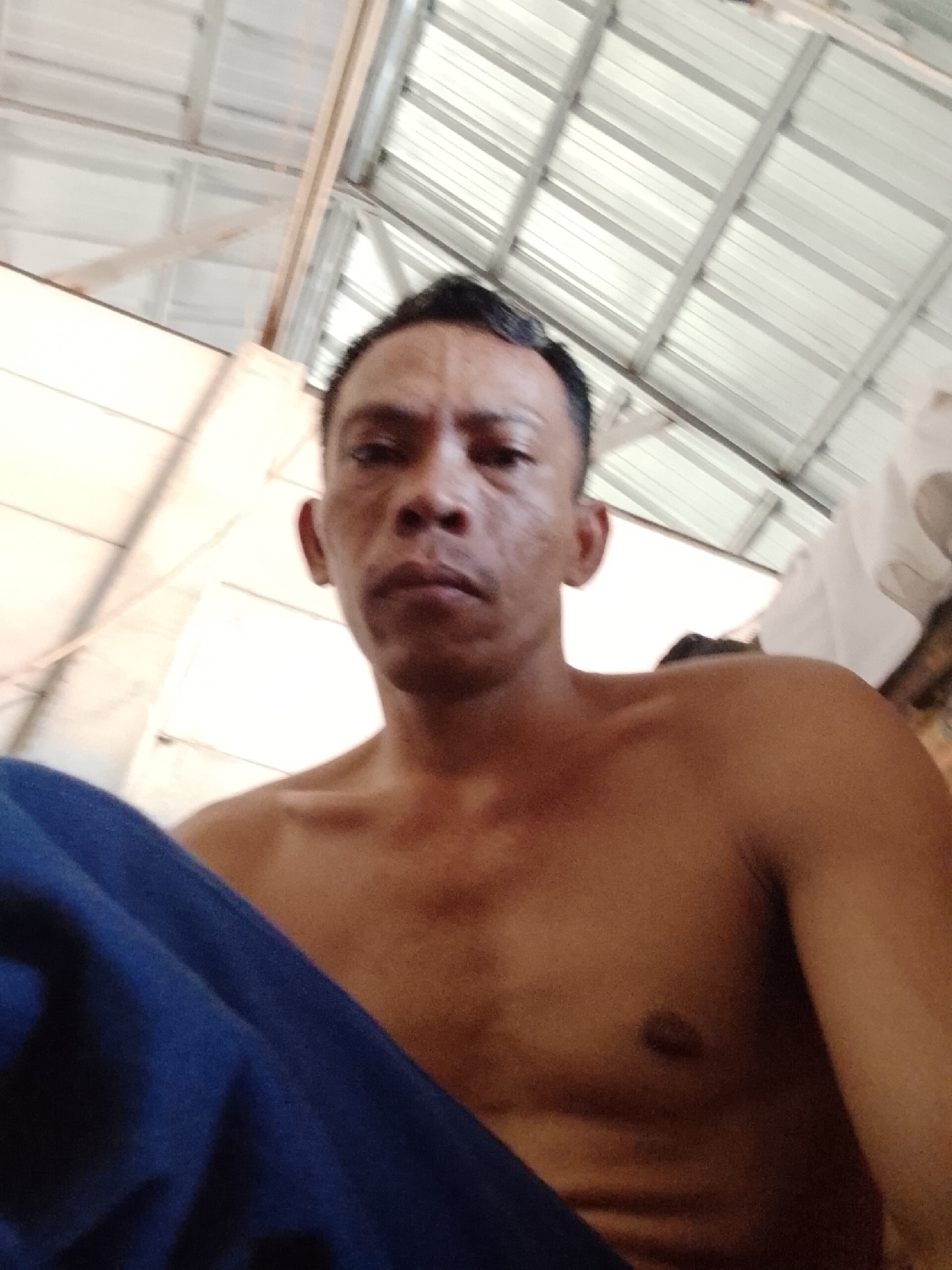 A shirtless man is sitting and looking at the camera. The location is Bukit Ketok, Indonesia. He is under a corrugated metal roof structure. A blue cloth covers part of his body.