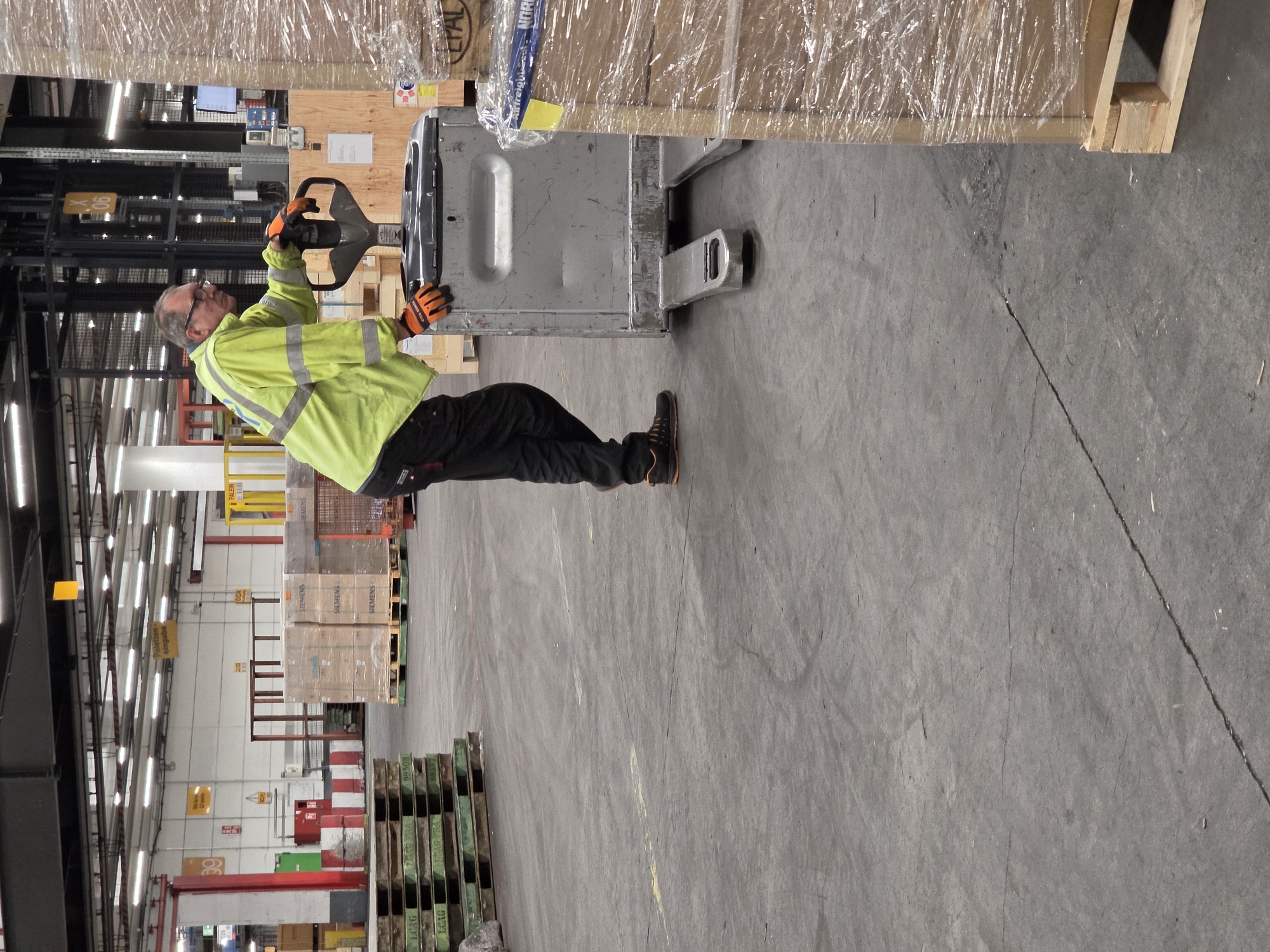 The image depicts a man wearing a bright yellow high-visibility jacket and black trousers, along with orange gloves and safety shoes, operating a silver pallet jack in what appears to be a warehouse environment. He is positioned on the right side of the frame, facing left, with his hands on the pallet jack's handle. The pallet jack is positioned next to a large stack of boxes wrapped in plastic, which are on a wooden pallet. In the background, there are more stacks of boxes on pallets, some with prominent "SIEMENS" branding, and industrial shelving units. The floor is concrete, and the ceiling is lined with fluorescent lighting. Signs in German are visible in the background, suggesting the location is in Germany, likely Frankfurt, given the context. One sign visible reads "Paletten eingabe" which translates to "Pallet input".