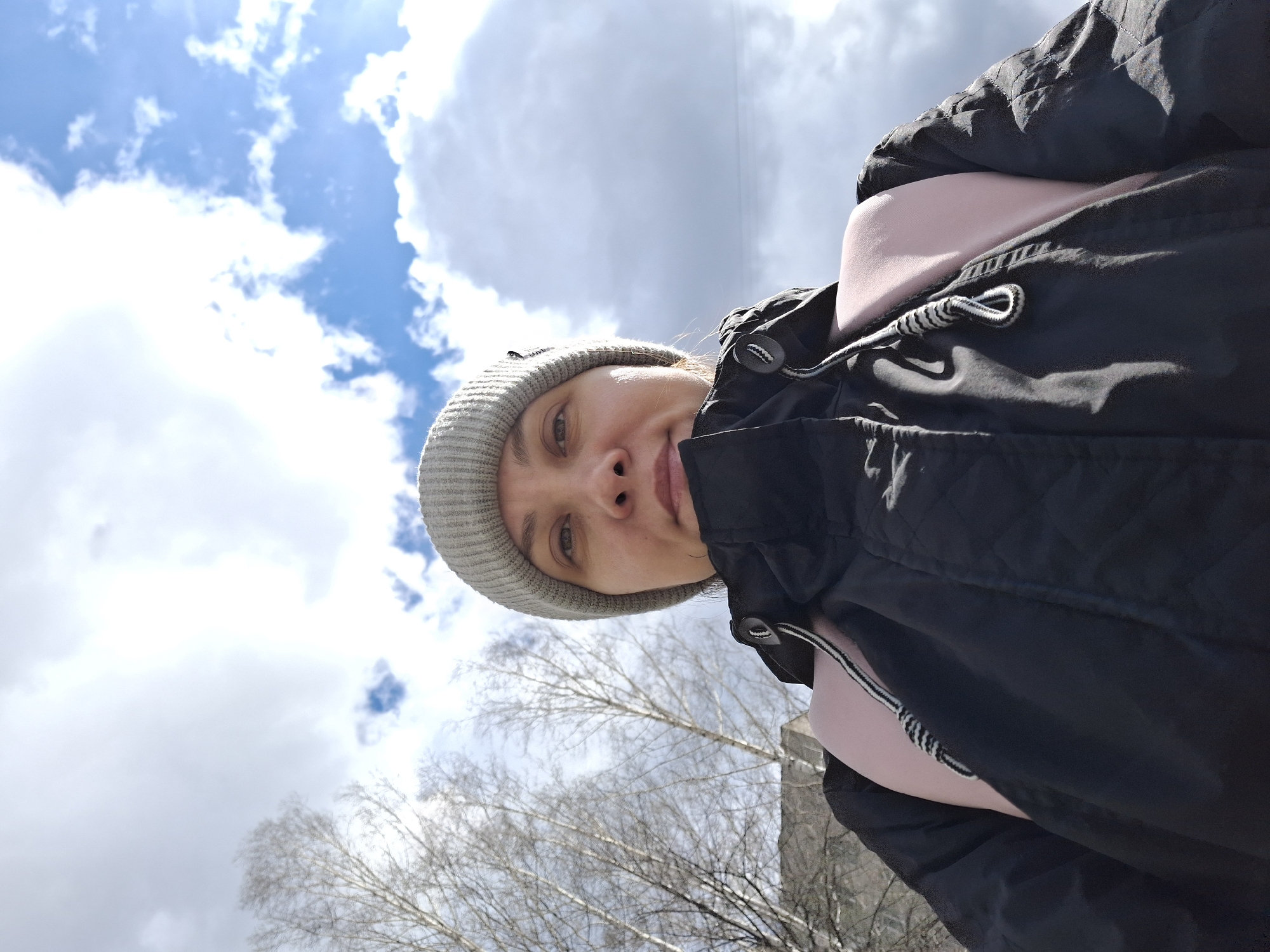 The image is a vertical, low-angle selfie taken outdoors, featuring a person against a bright, cloudy sky. The subject, likely a woman, is positioned in the right half of the frame, looking directly into the camera with a gentle, subtle smile. She is wearing a light grey knit beanie and a black quilted jacket. A light pink garment, possibly a top or a backpack strap, is visible beneath the jacket.
The background is dominated by a clear blue sky interspersed with numerous white cumulus clouds. A bright light source, likely the sun, is present in the upper left, causing a lens flare effect. In the lower left quadrant of the image, the intricate, bare branches of deciduous trees are visible, silhouetted against the sky, indicating a late winter or early spring season. A faint outline of a grey building or structure can be seen on the far right.
The photo captures a casual moment outdoors during the daytime. The attire suggests cool weather. The visible elements combine to depict an outdoor scene in Chaikovsky, Russia, characterized by bright daylight and early spring or late winter conditions. No visible text is present in the image.