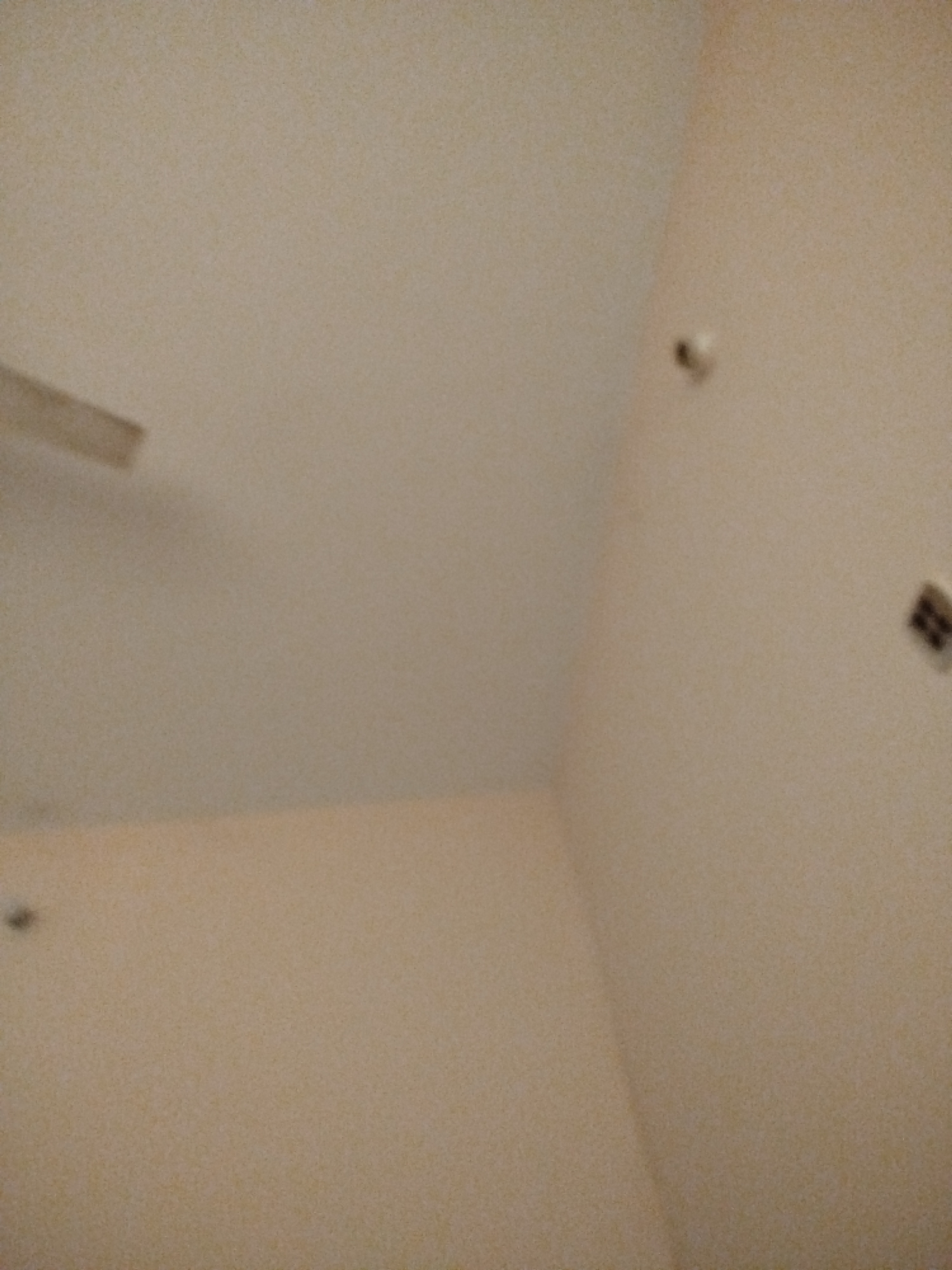 The image depicts a ceiling in what appears to be an indoor space. There is a partial view of a ceiling fan blade on the left side of the frame. On the wall, there are two small, dark circular objects that could be fixtures or imperfections. In the upper right corner, a dark, rectangular object is visible on the wall. The walls and ceiling are a light, neutral color, possibly beige or off-white. The lighting in the room is dim, contributing to the somewhat blurry appearance of the scene. The image is taken from a low angle, looking upwards. There are no people or any discernable actions occurring within the frame. The location in the city of Lassan, Pakistan, cannot be determined from the visual information provided in the image.