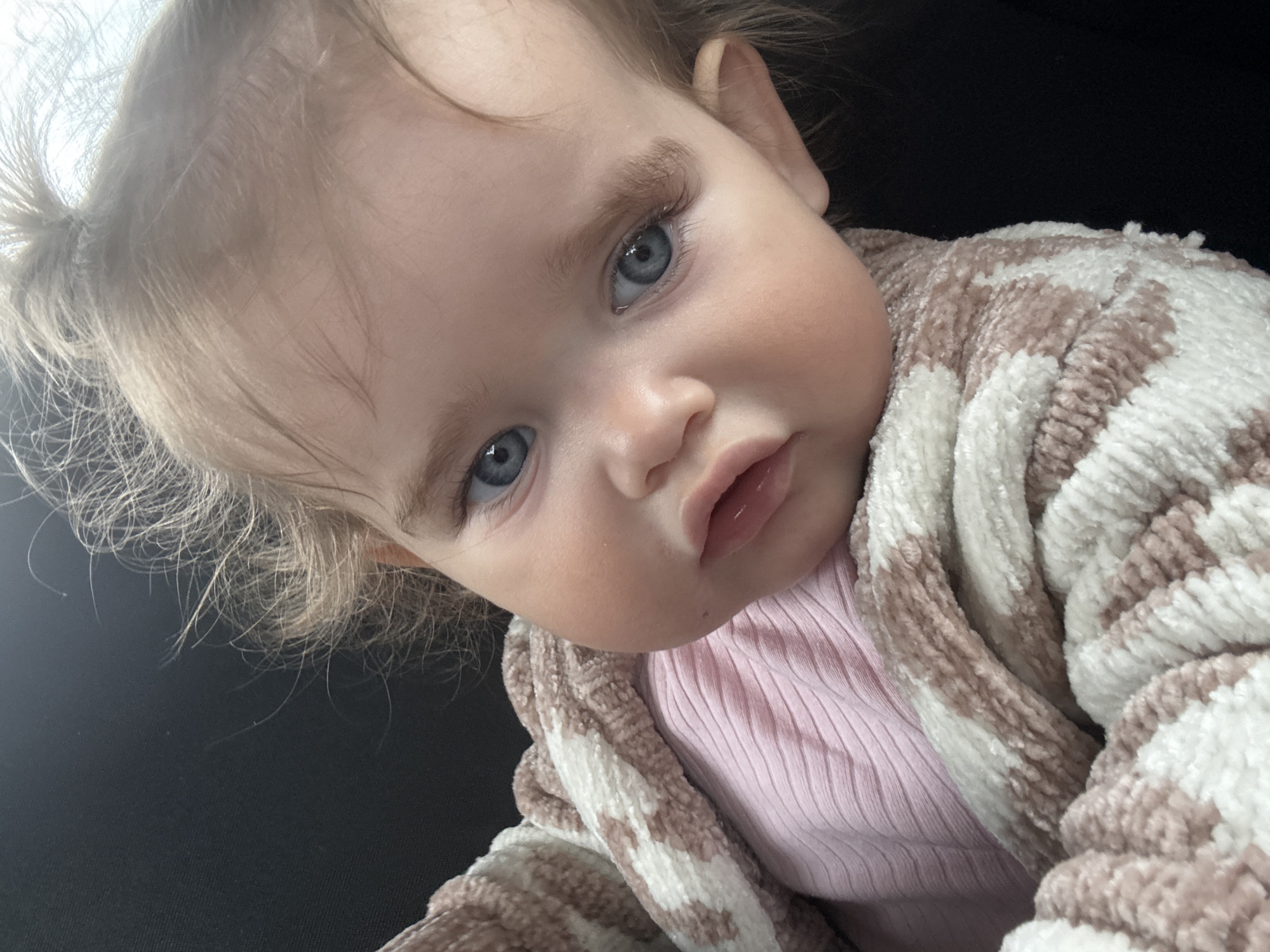 A close-up, head-and-shoulders shot of a baby girl with blonde hair and bright blue eyes is featured. She is wearing a pink ribbed shirt and is partially wrapped in a fuzzy blanket with a pink and white striped pattern. The baby's head is tilted, and her gaze is directed slightly off-camera. The background is dark and indistinct, suggesting an indoor setting. There is no discernible information indicating a location in Braunfels, Germany.