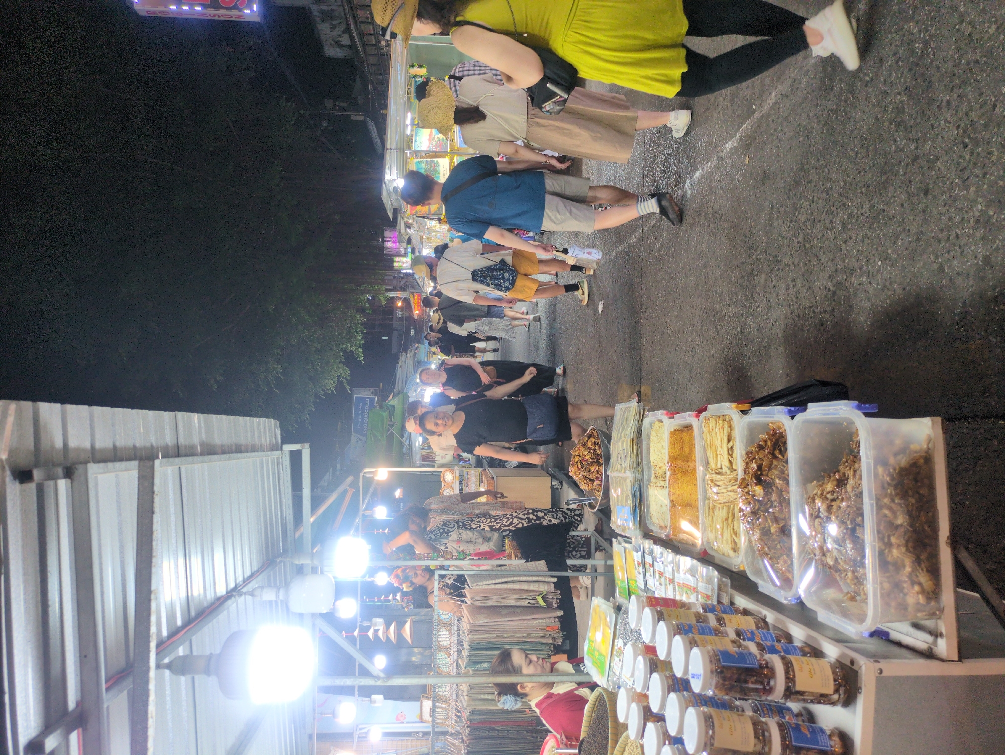 The image shows a night scene at a street market in Phú Quốc, Vietnam. People stroll along a narrow paved street, browsing stalls illuminated by bright lights. One stall features various dried foods in clear plastic containers and jars. Another stall displays clothing items. The atmosphere is lively, with many people present.