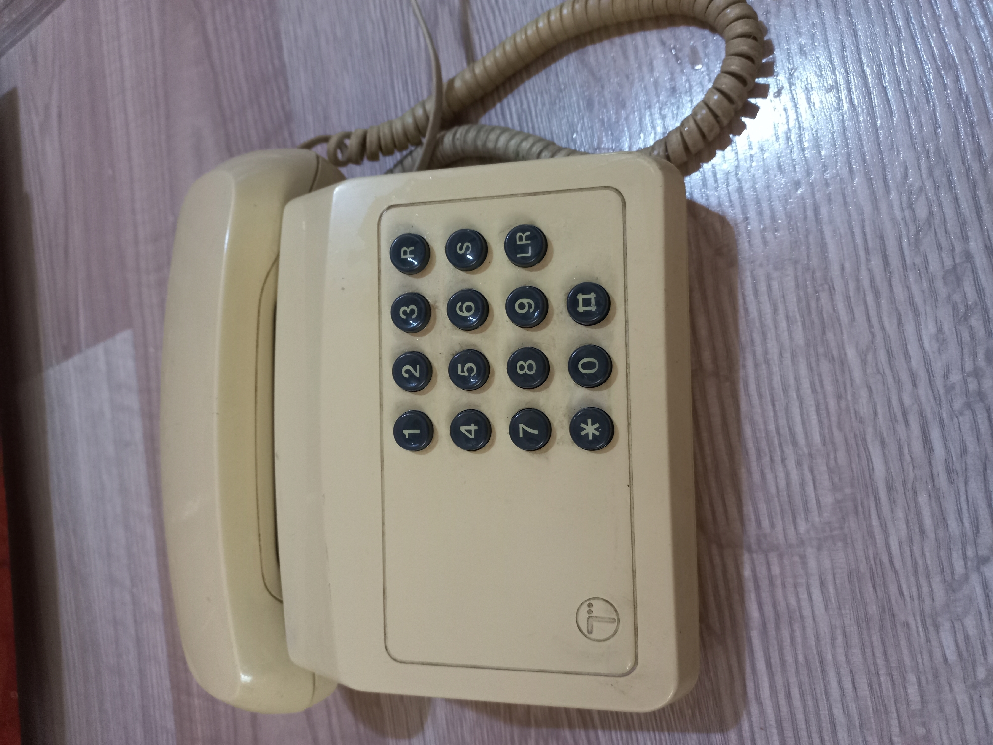 A vintage, cream-colored rotary dial telephone with a coiled cord rests on a light-colored, wood-grain linoleum floor. The telephone features a keypad with numbers 0-9, as well as buttons labeled R, S, and LR. A circular logo with a stylized "T" is visible on the left side of the keypad. The telephone is positioned at an angle, with the handset resting on top of the base. The overall scene is dimly lit, with some reflections visible on the floor. The image does not provide any specific clues to its location within the city of Ashgabat, Turkmenistan.