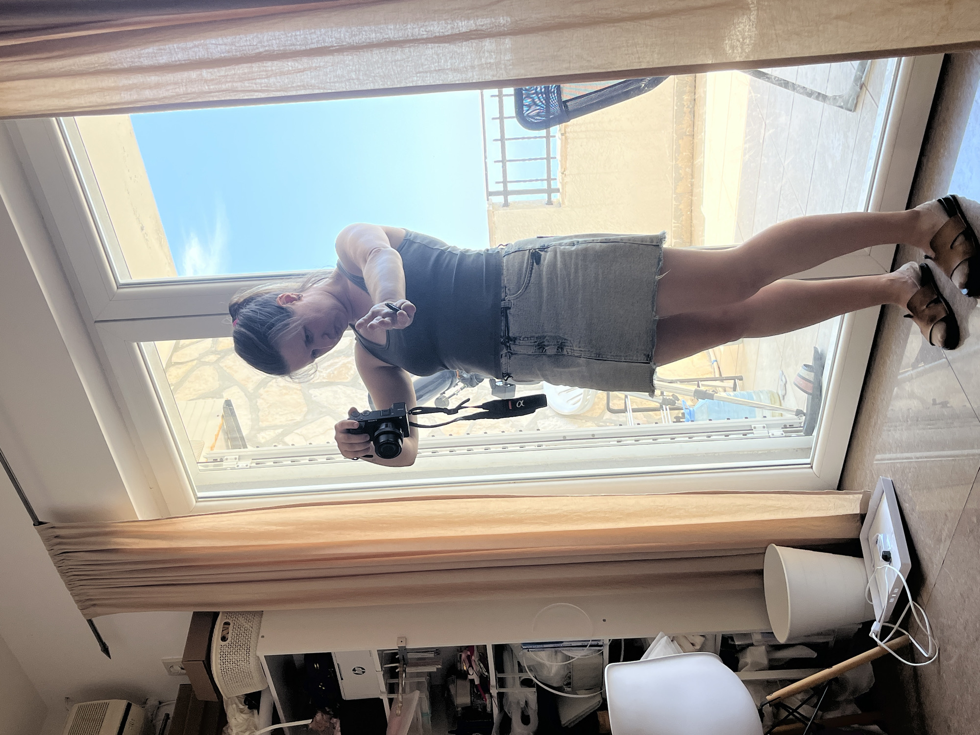 A woman stands indoors, near a large sliding glass door, holding a camera and looking at its screen. She is wearing a dark tank top and a light denim skirt. Her hair is pulled back in a ponytail. The background visible through the glass door shows a balcony with a railing, a blue sky, and some distant buildings. Inside, to the left of the woman, is a white chair, a white trash can, and a white tray with a charging cable. To the left of the door, a beige curtain hangs from a rod. The floor is tiled. The scene appears to be located on a balcony or terrace in Budva, Montenegro, evidenced by the architecture and the view of the sea.