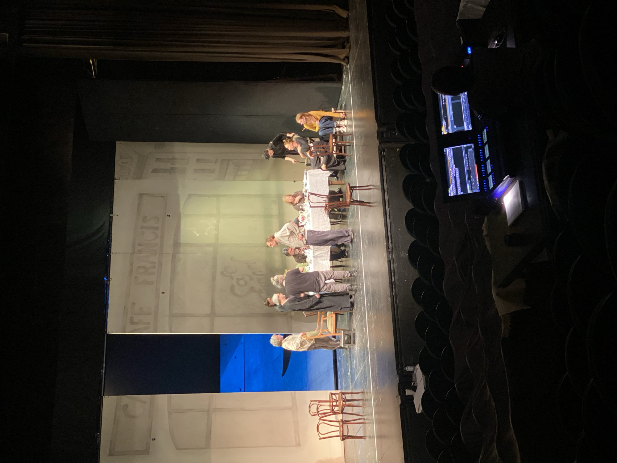 The image shows a theater production on stage. The backdrop is a painted facade of "Cafe Francis". A long table covered with a white tablecloth is center stage, surrounded by actors. Some actors are seated at the table, while others are standing or interacting with the set and props. Additional props include chairs and a blue screen. The image is taken from the audience, showing rows of seats. An individual is to the right, operating a control panel with screens displaying production software. The location, Khimki, Russia, is not explicitly evident from the image itself.
