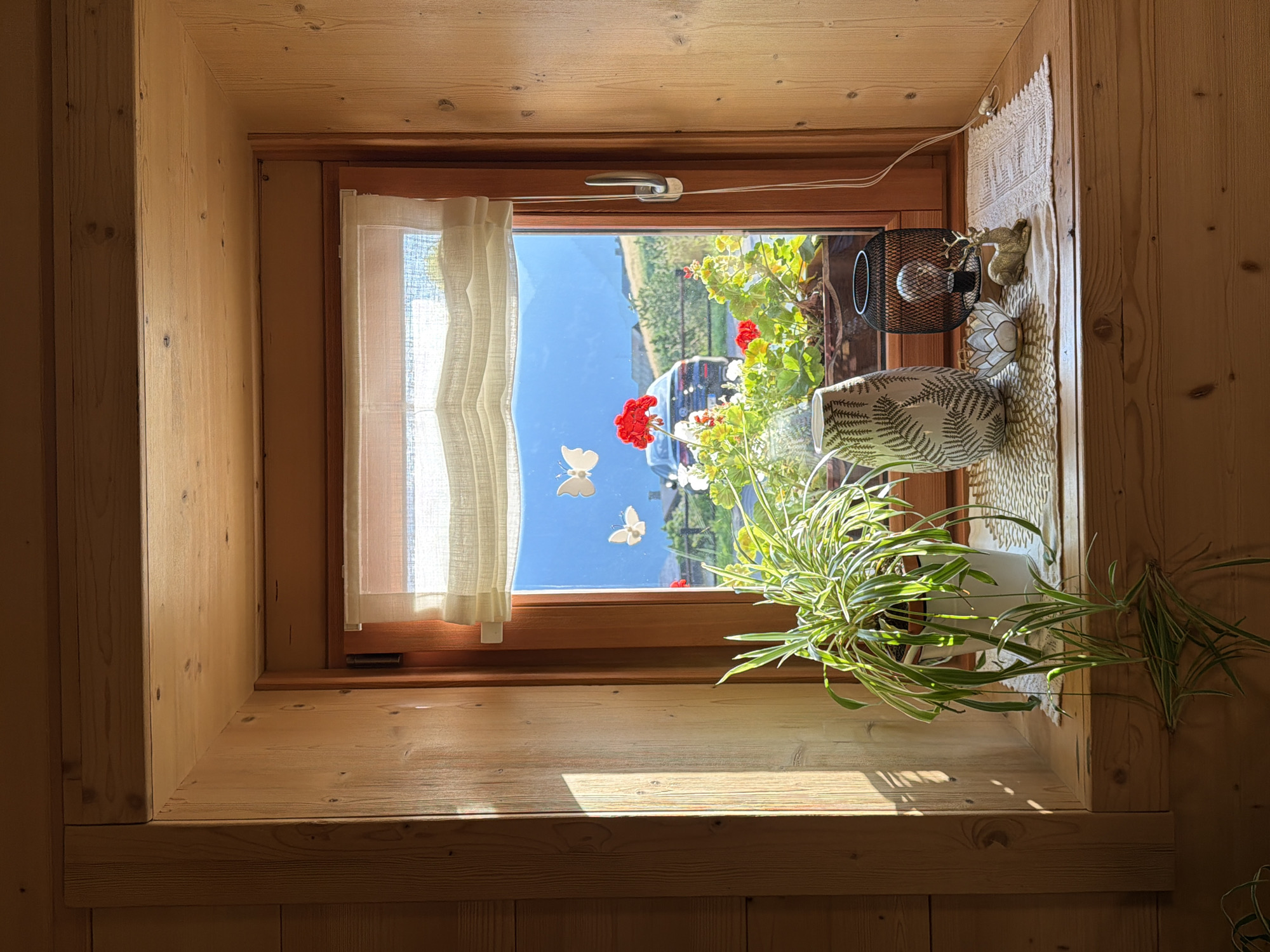 The image displays a sunlit wooden windowsill in Sauris/Zahre, Italy, featuring a Spider Plant in a white pot, a decorative vase with a fern pattern, a small lotus-shaped candle holder, a black decorative lantern with a light bulb, and a golden deer figurine. Beyond the window, a bright blue sky is visible, with a scenic view of green hills, a white car, and red flowers in planters. Two white butterfly decals are affixed to the windowpane. The wooden interior of the window frame and the surrounding wall are visible, showcasing the natural grain of the wood.