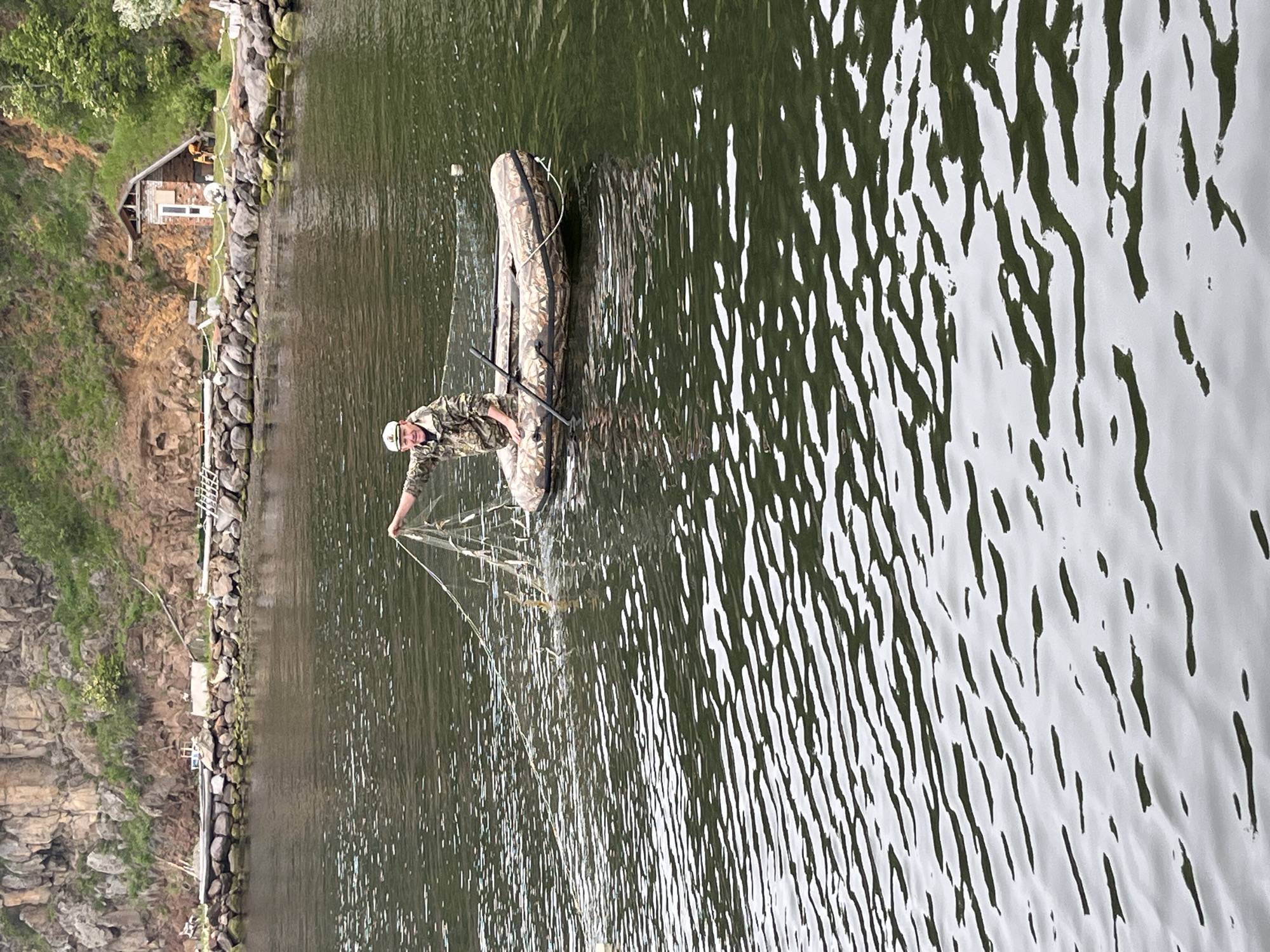 A person in camouflage clothing and a white cap is in an inflatable boat on the water, holding a fishing net with fish. The boat is camouflaged and has oars. In the background, there is a rocky cliff with green vegetation. Along the shoreline, there is a stone wall, and a small wooden building with a red brick chimney. The water is choppy, with reflections of the sky and surrounding environment. The location is the Bay of Sovetskaya Gavan', Russia.