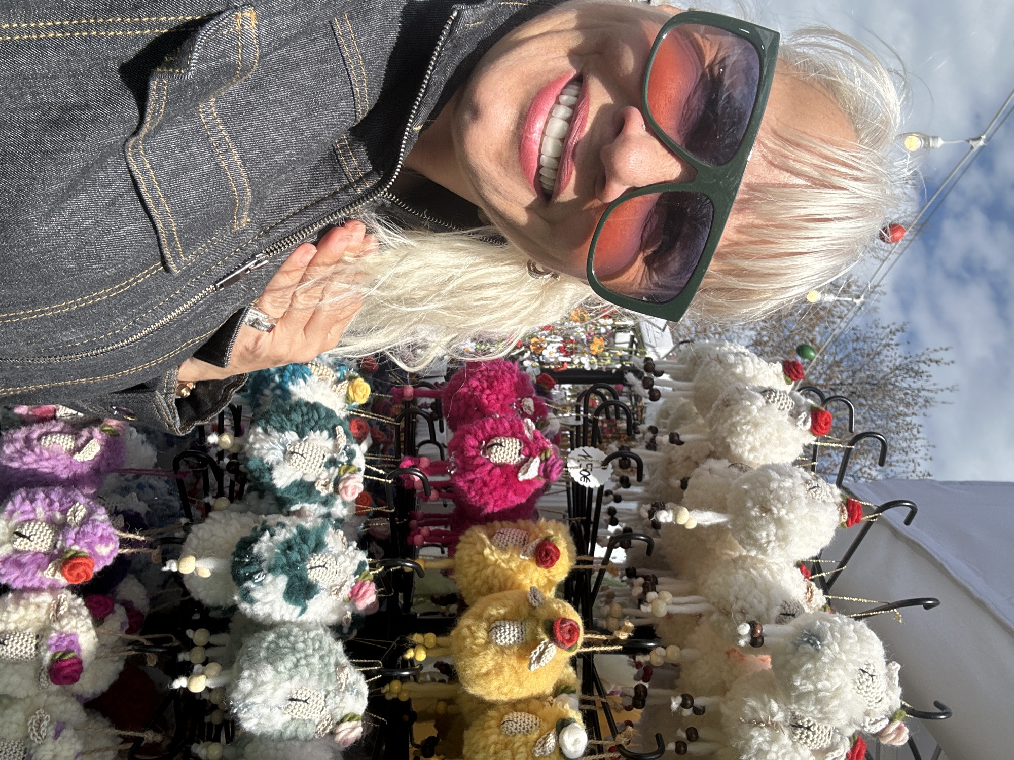 A woman with blonde hair and sunglasses smiles at the camera while standing behind a display of handcrafted ornaments. The ornaments appear to be small, plush animals with embroidered faces and floral decorations, hung on black metal hooks. The colors of the ornaments include purple, green, pink, yellow, and white. The woman is wearing a denim jacket. The sky is visible in the background. Based on the presence of handcrafted items displayed outdoors and a white tent partially visible on the right, the location could be a craft fair or open-air market. Given the request for the location, this could be within the city of Sofia, Bulgaria.