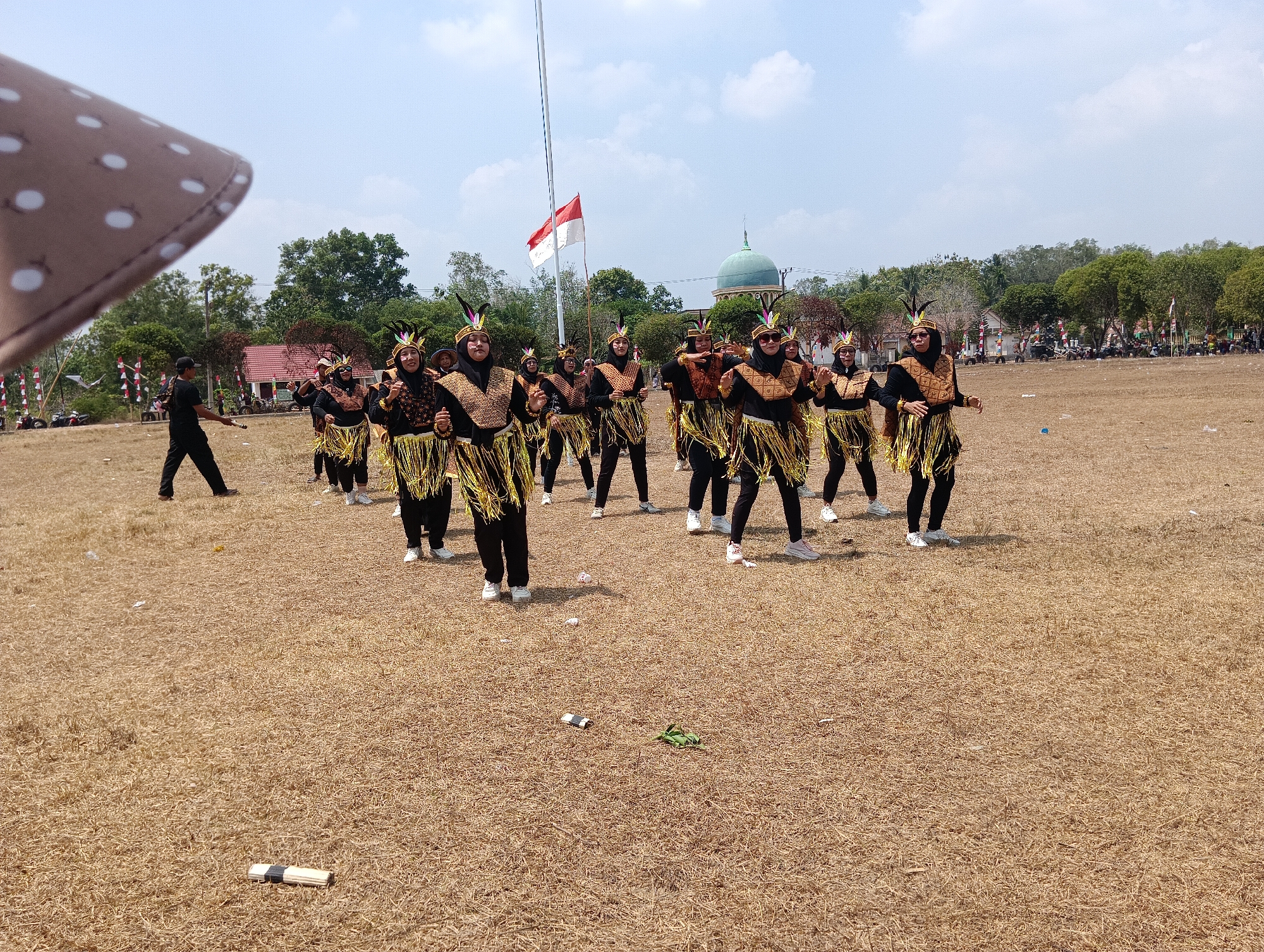 A group of approximately 12 individuals dressed in ceremonial costumes and headdresses are performing a dance on a dry, grassy field under a partly cloudy sky. The costumes consist of black tops and bottoms, adorned with gold fringe skirts and decorative gold chest pieces. Some performers are wearing face paint. In the background, a flagpole with the Indonesian flag is visible, along with trees, buildings, and a dome-shaped structure. A person in dark clothing is walking away from the main group on the left side of the frame. The ground is littered with some debris, including a white tube-like object and a green plastic item. The scene appears to be an outdoor event in Pematang Jaya, Indonesia.