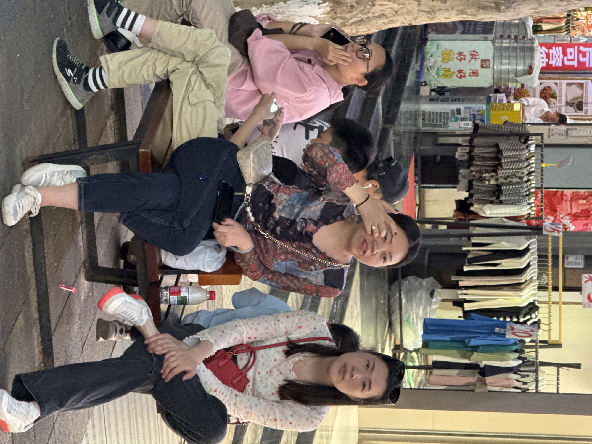 The image depicts a scene on a street in Beibei, China, where a group of individuals are sitting and interacting. Three women are prominently featured in the foreground. One is wearing a white shirt with colored polka dots, dark pants, and white sneakers with orange accents. She has a red handbag slung over her shoulder and sunglasses perched on her head. Another woman is wearing a patterned blouse and jeans. The third woman has a pink shirt and khaki pants. All three are seemingly engaged in conversation and using mobile phones.
Behind the women, there is a commercial area visible, with racks of clothing displayed. There are various signs and displays with Chinese characters, and a man in a white shirt.
The scene appears to be a candid moment captured on a busy street in the city.