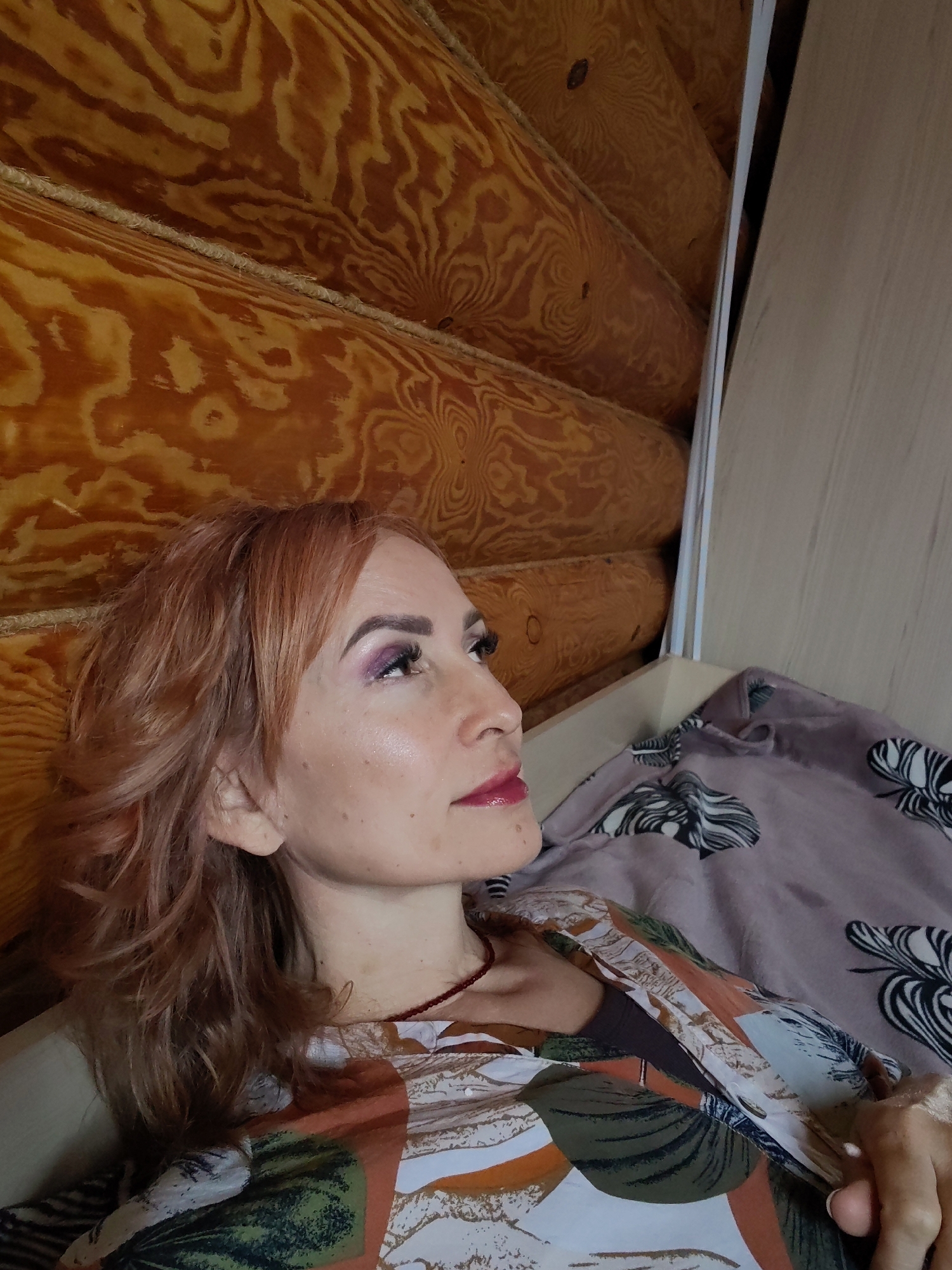 A fair-skinned woman with reddish-blonde, wavy hair is seen in a close-up, side-profile shot. She has dark, heavily mascaraed eyelashes and purple eyeshadow. She is wearing a patterned top with hues of green, brown, white, and orange. A thin, red necklace is visible around her neck. Her lips are glossy and red.
She is lying in a bed with grey-toned bedding that has a leaf-like pattern. Behind her, the wall is constructed of interlocking, polished wood logs, typical of a log cabin. To her right, there is a light-colored, flat surface, possibly a piece of furniture or part of the wall.
The location is Старое Михайловское, Russia, as inferred from the image description. The setting appears to be indoors within a rustic, possibly rural, home.
