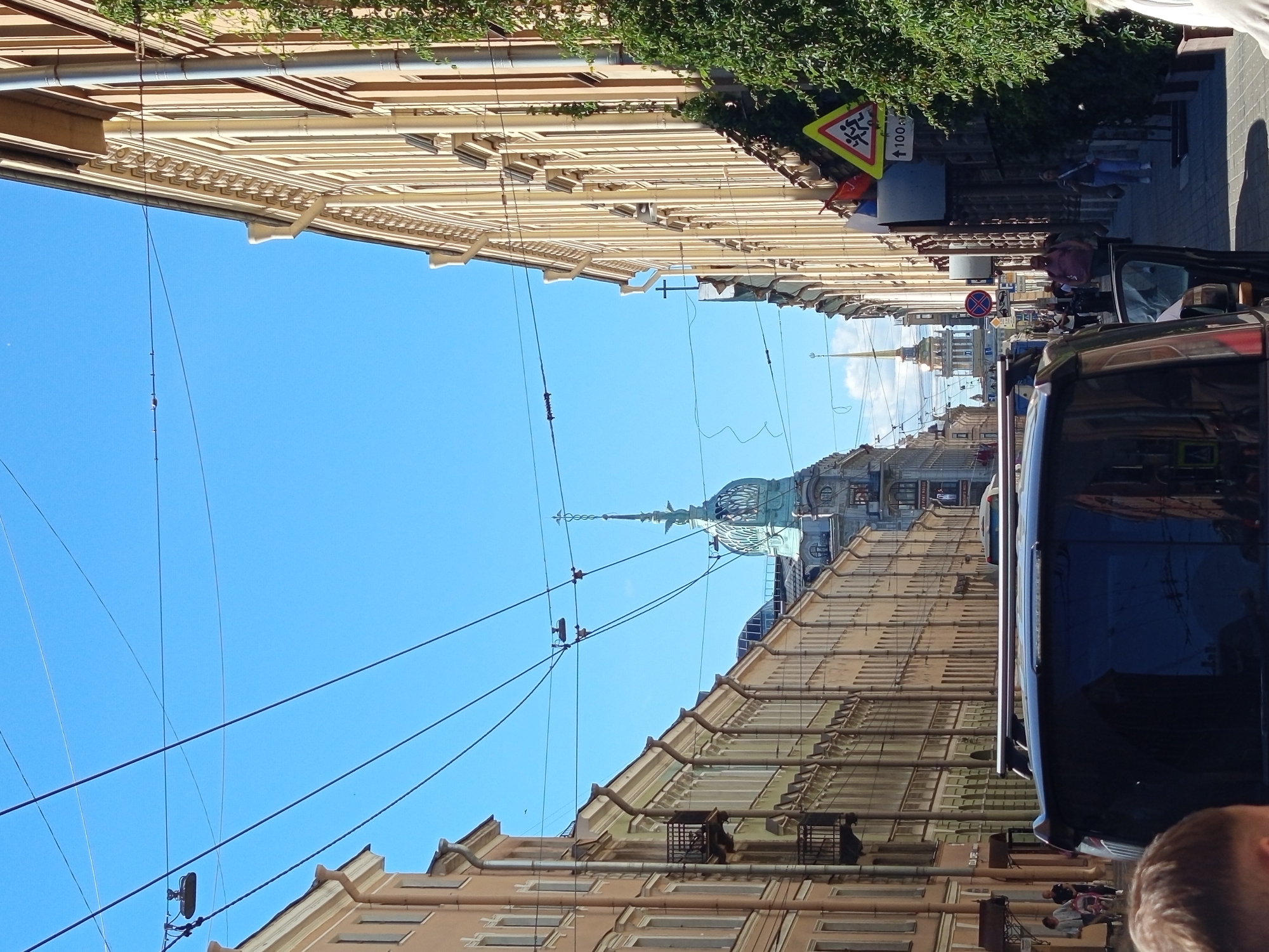 The image depicts a street scene in Saint Petersburg, Russia, likely on a sunny day with a clear blue sky. The perspective is from behind a dark-colored SUV, looking down a street lined with multi-story buildings. The architecture of the buildings suggests a historical European city, with ornate facades, arched windows, and decorative cornices. Electrical wires crisscross the sky, typical of older urban infrastructure.
On the right side of the street, a green, leafy tree provides a natural element against the buildings. A yellow triangular sign with a symbol of running figures indicates a pedestrian crossing, with the number "100" below it, possibly denoting a distance or speed limit. Further down the street, a prominent steeple with a gold spire rises above the rooftops, hinting at a significant landmark, possibly a church or administrative building.
Several people are visible on the sidewalk, walking in various directions. On the left side, a couple is walking with a stroller. On the right, individuals are seen strolling along the pedestrian path. The foreground is dominated by the rear of the SUV, with its rear window reflecting the surrounding buildings and sky. A person's head is visible in the bottom left corner, slightly out of focus, suggesting the photographer is positioned behind this vehicle. The overall impression is a lively urban environment with a blend of historic architecture and everyday city life.
