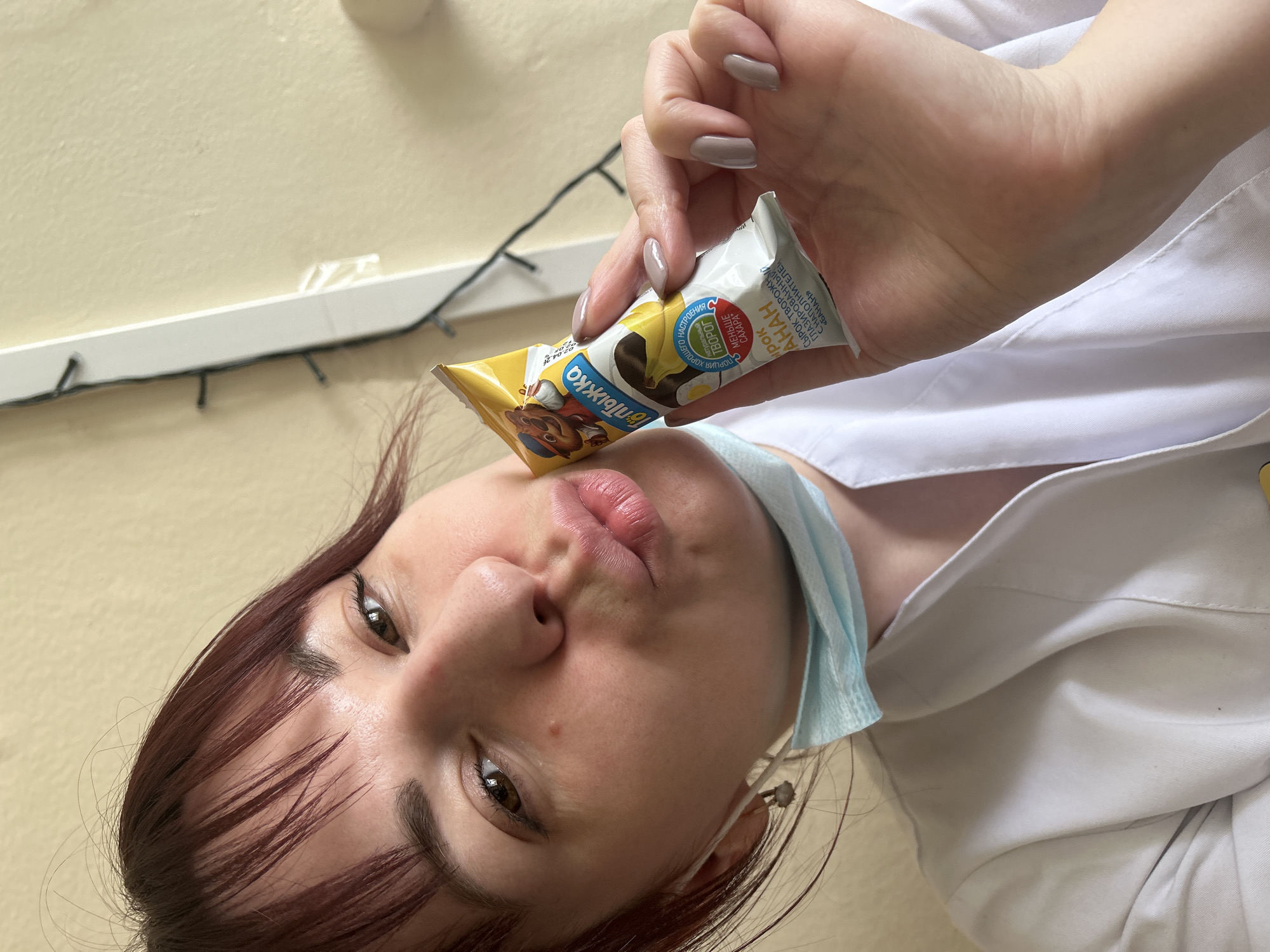 A woman wearing a white medical coat and a surgical mask around her neck holds a "Топтыжка" ("Toptyzhka") brand chocolate-covered cheese curd bar with banana flavor in her right hand, positioning the bar near her face. The packaging indicates the product contains less sugar and natural tvorog (a type of quark cheese). The background appears to be an indoor setting, with string lights visible in the top left corner. Although the city Фоки is mentioned, the location cannot be verified in the image.