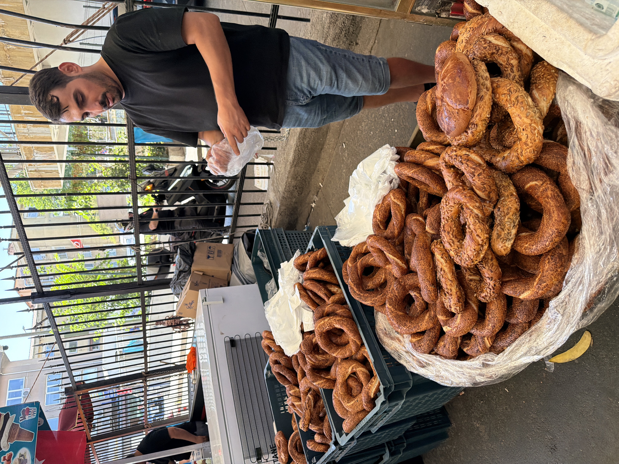 The image depicts a street vendor in Kadıköy, Turkey, preparing to package simit, a type of Turkish bagel, for a customer. A man in a black t-shirt and denim shorts stands behind a stall laden with simit. He is holding a clear plastic bag in his hands, seemingly preparing to place simit into it. In the background, a metal fence separates the vendor's stall from the street. Several crates overflowing with simit are visible, along with a white freezer and some cardboard boxes. A motorcycle is parked behind the fence. The scene is set outdoors on a paved street, typical of a bustling market area. The overall impression is of a lively, everyday street scene in a Turkish city.