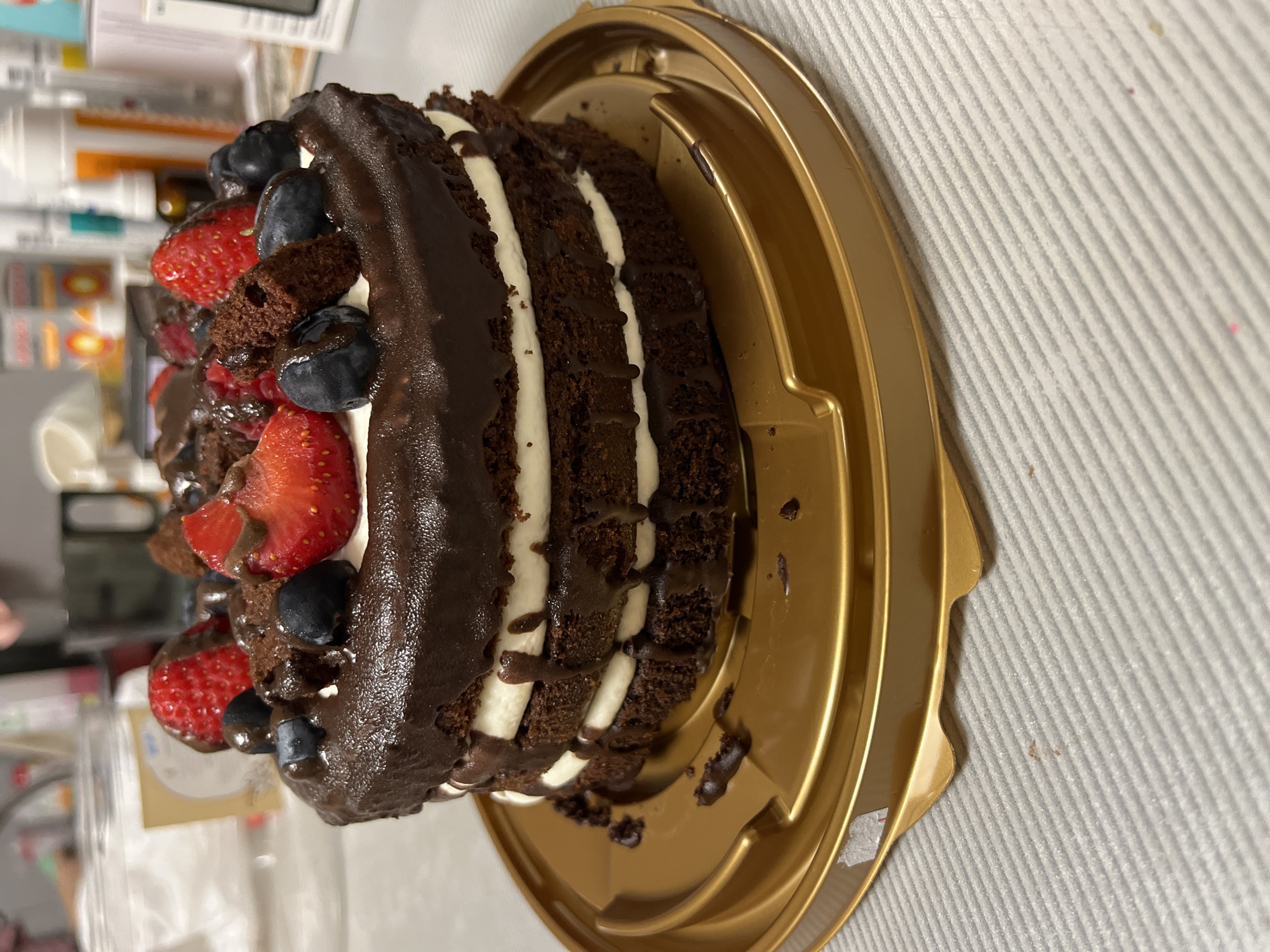 A three-tiered chocolate cake, topped with fresh strawberries, blueberries, and chocolate pieces, sits on a golden cake stand. The cake is decorated with white cream and dark chocolate frosting that drips down the sides. The stand is placed on a white, ribbed tablecloth. The background is blurred and shows a shelf with various bottles and containers, suggesting an indoor setting. There is no indication of the city of Sochi, Russia, or any specific location.