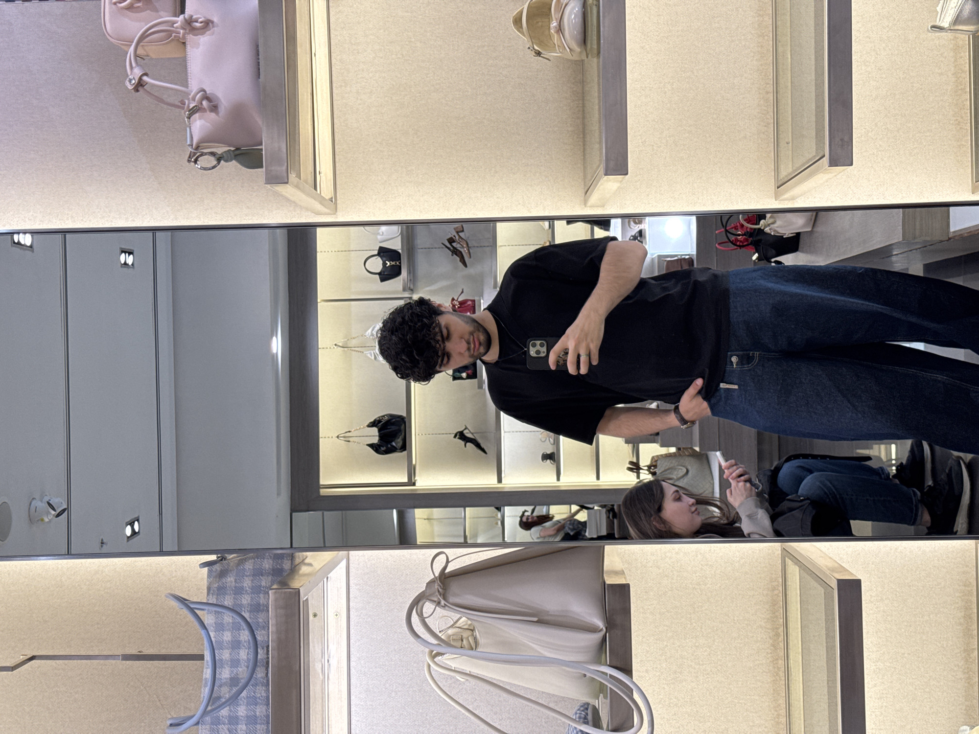 A man with curly hair, wearing a black t-shirt and jeans, is taking a selfie in front of a mirror. The mirror reflects a store with displays of handbags and other accessories. In the reflection, a woman is seated, looking at her phone. The location appears to be a retail store, possibly in Yerevan, Armenia.